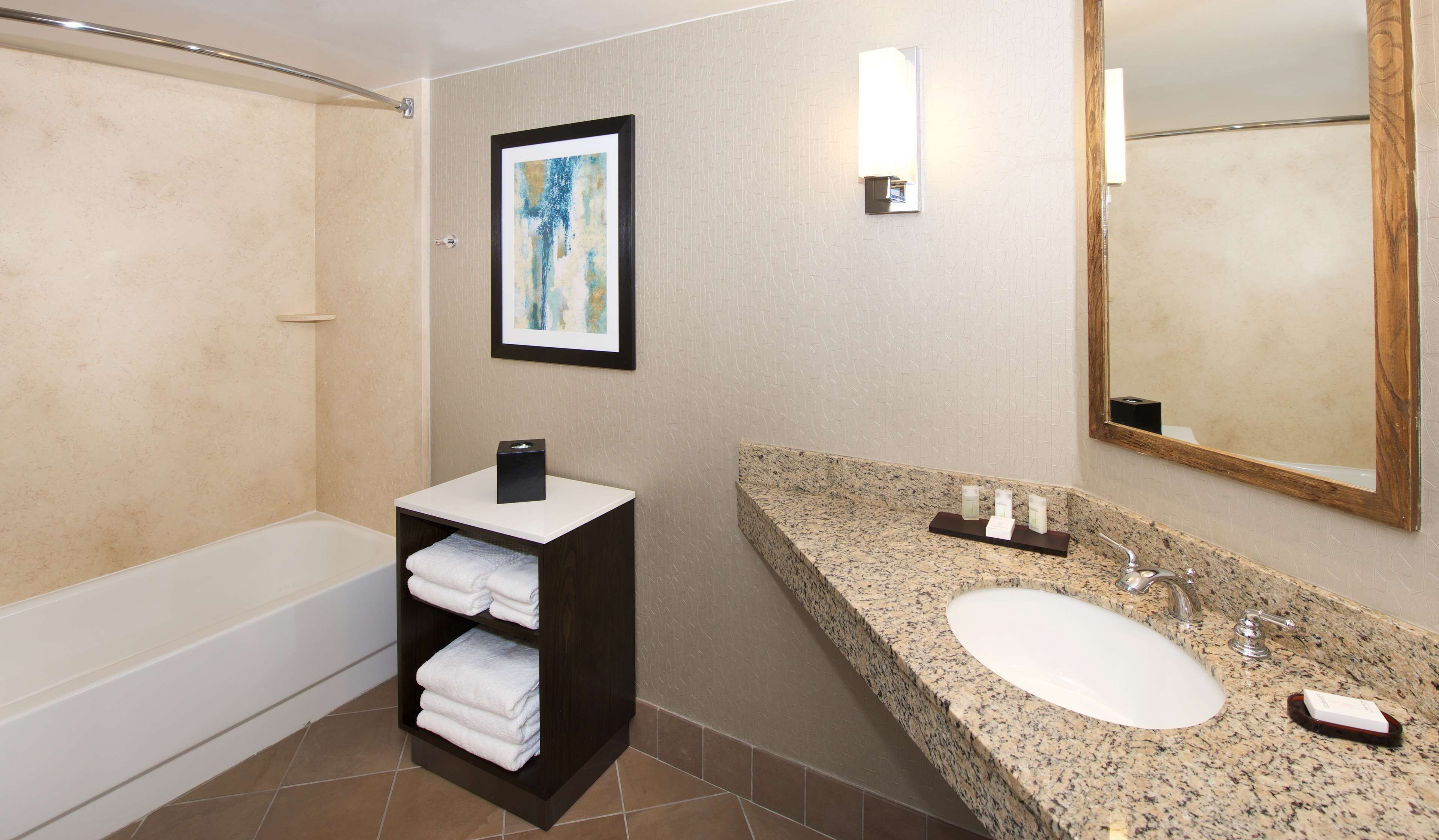 A modern bathroom with a bathtub, a vanity with a granite countertop, a sink, and a large mirror. There is a framed artwork above the vanity and a towel rack with folded towels.