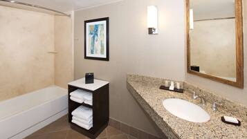 A modern bathroom with a bathtub, a vanity with a granite countertop, a sink, and a large mirror. There is a framed artwork above the vanity and a towel rack with folded towels.