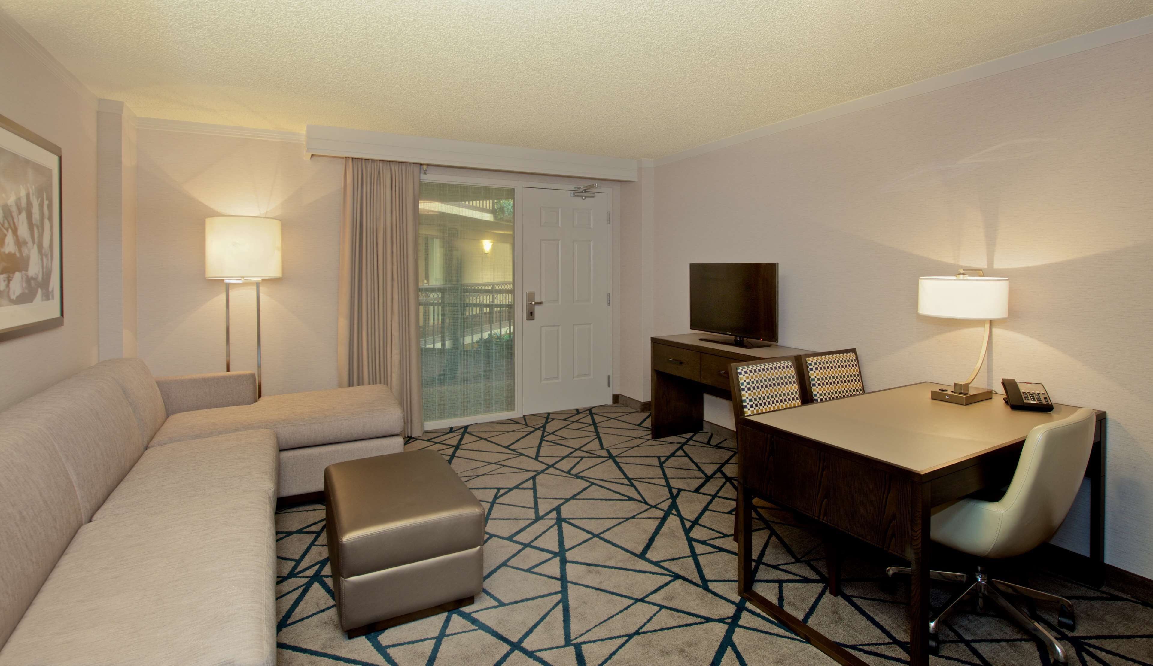 A hotel room with a sofa, ottoman, a desk with a chair, a TV, and a glass door leading to a balcony.
