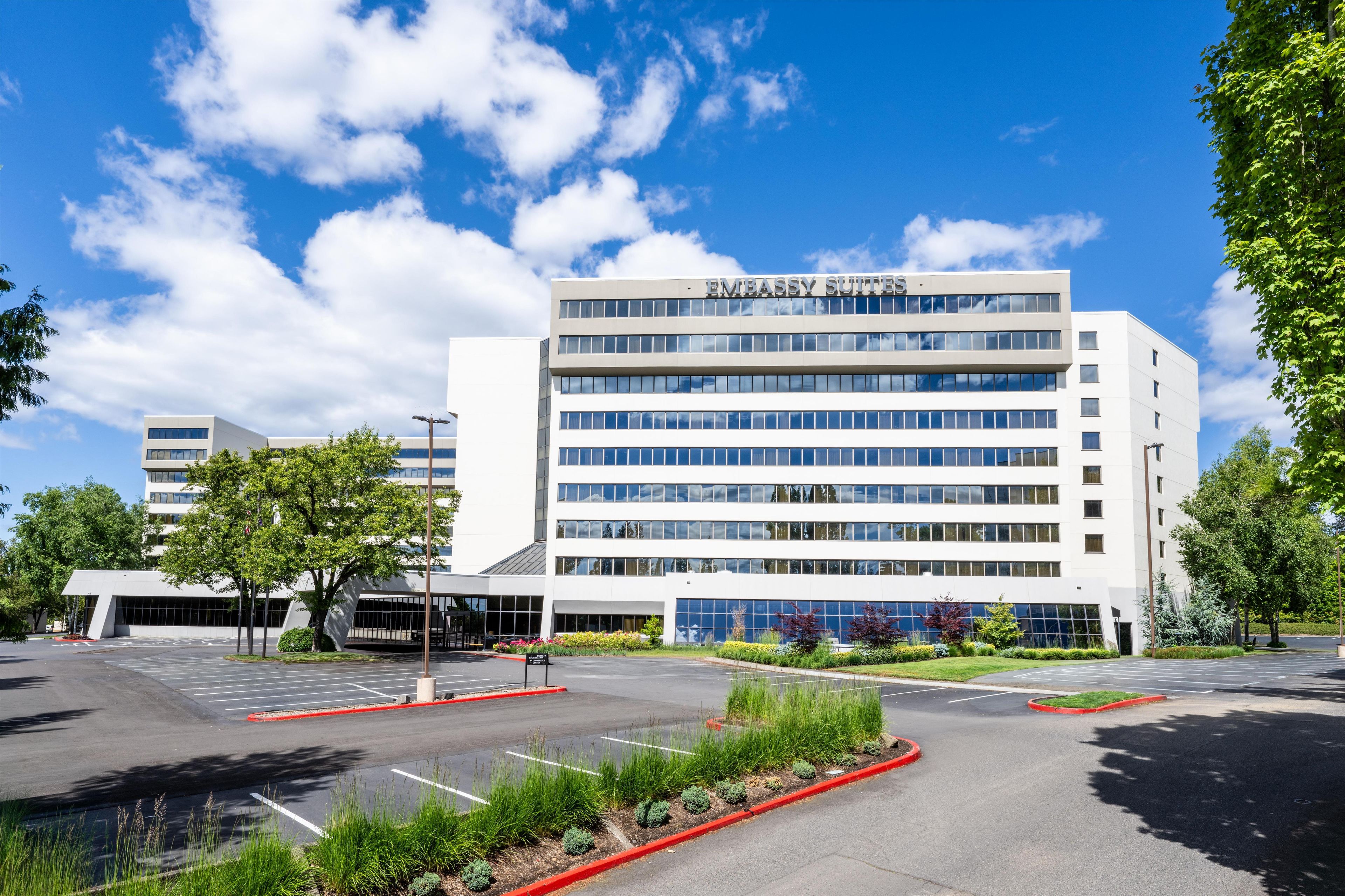 A modern building with 'EMBASSY SUITES' signage, a parking lot, and surrounding greenery.