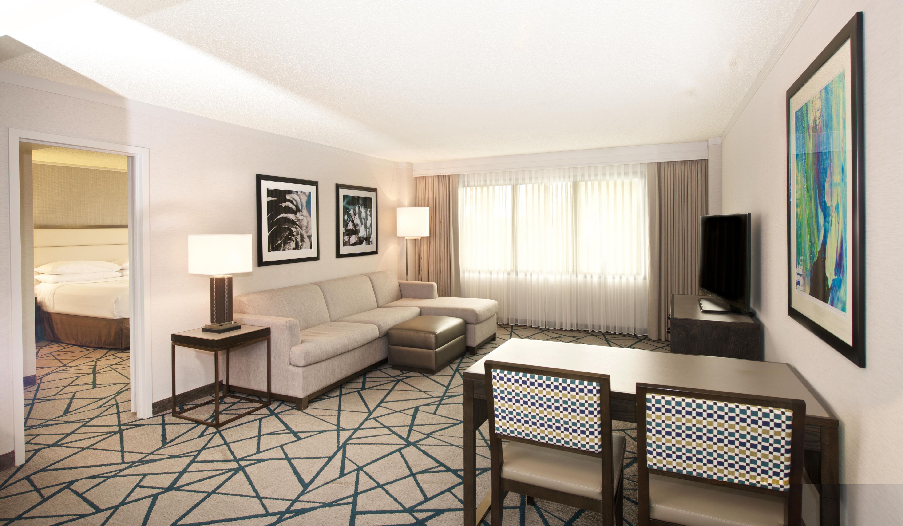 Habitación de hotel con cama, sofá, televisor, escritorio con sillas y obras de arte en las paredes.