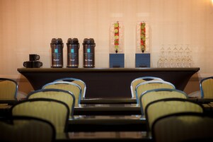 Zona de bar con una barra que muestra tres dispensadores de agua caliente, una máquina de café y un dispensador de bebidas. Detrás de la barra, hay dos dispensadores verticales de bebidas con guarniciones coloridas.
