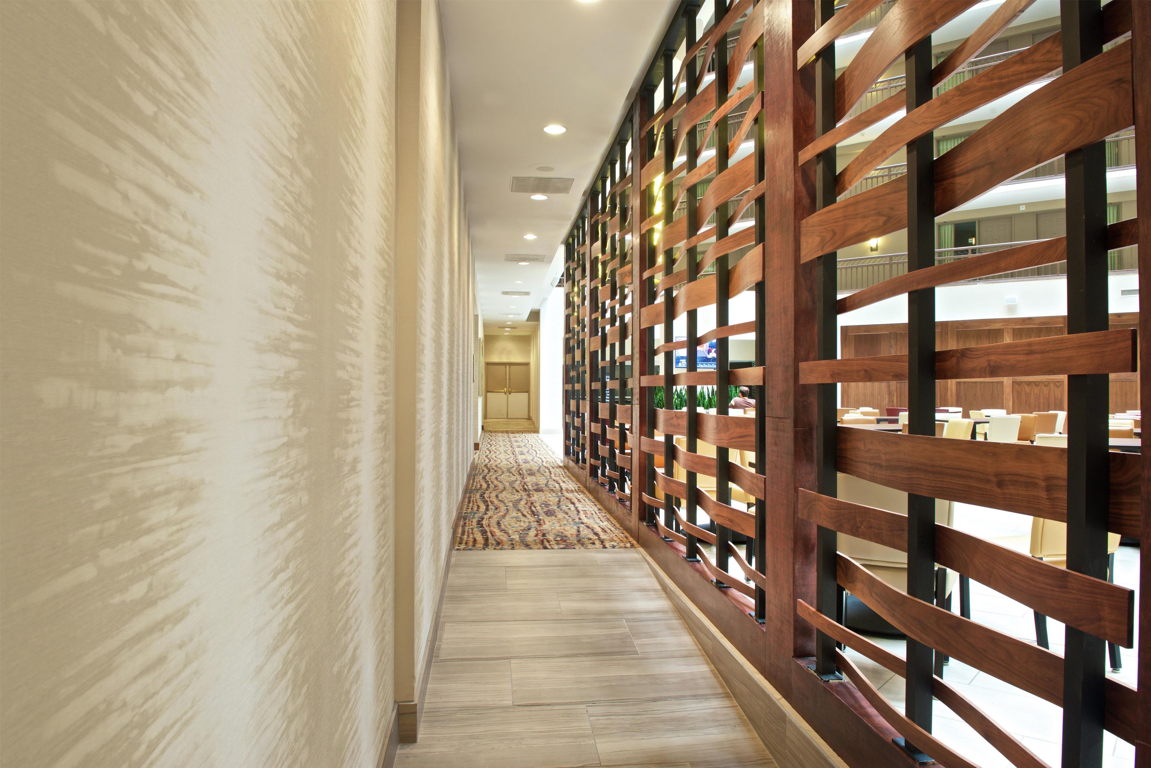 A corridor with patterned carpet, wooden paneling, and a view of a seating area with tables and chairs.