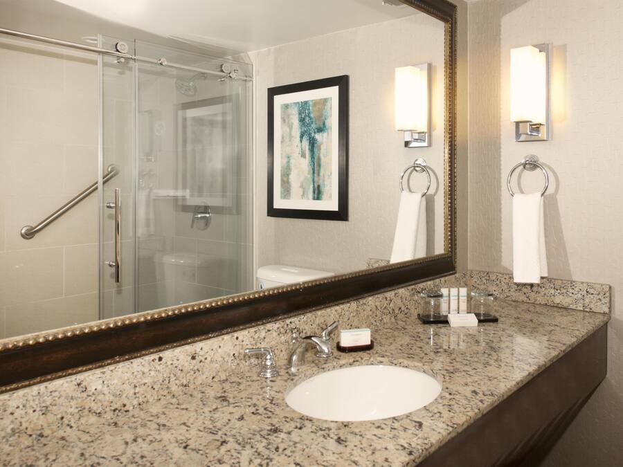 A bathroom with a glass shower enclosure, a sink with a granite countertop, and a large mirror with a decorative frame.