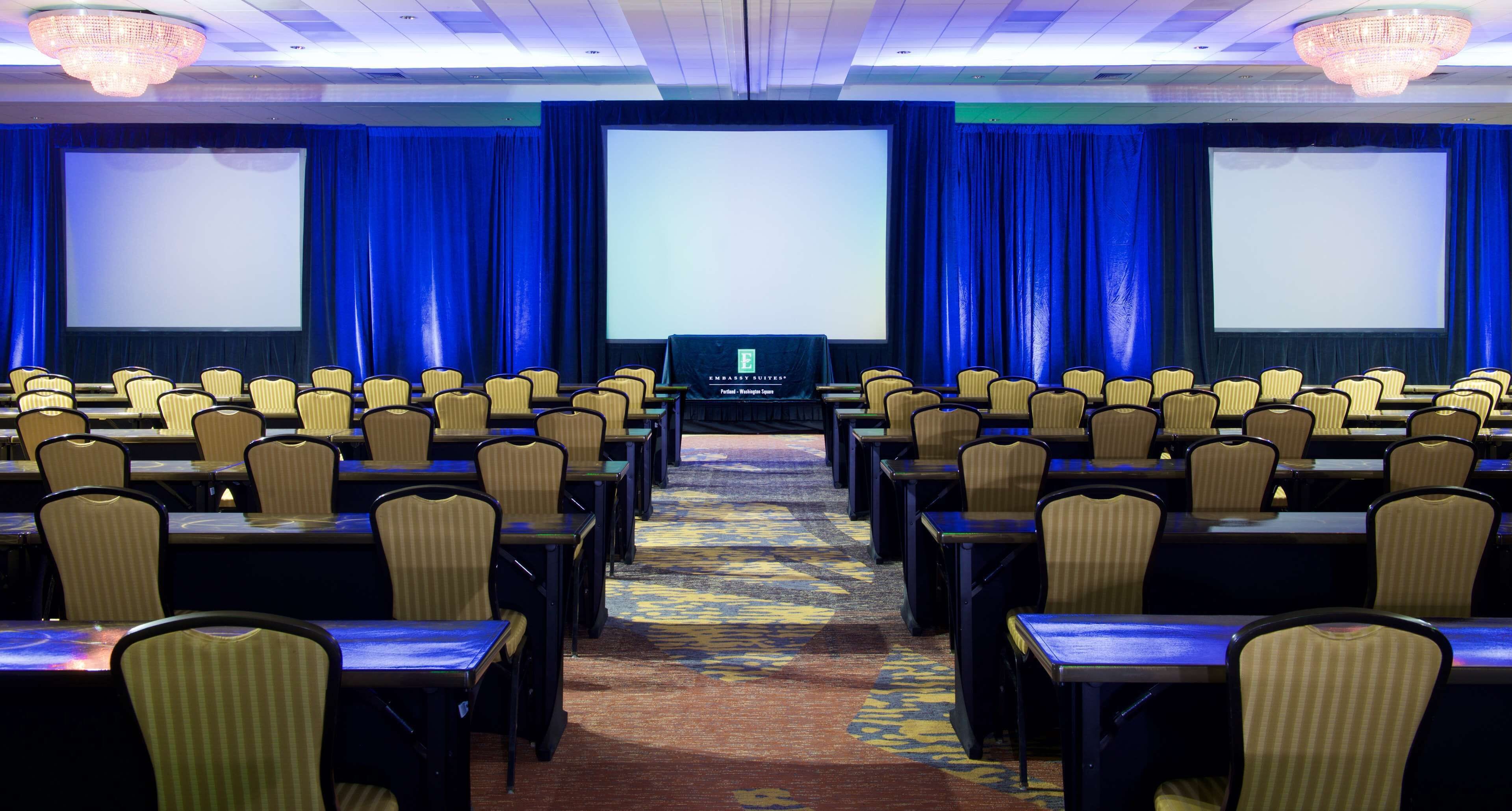A conference room with rows of tables and chairs, a large screen, and blue curtains.