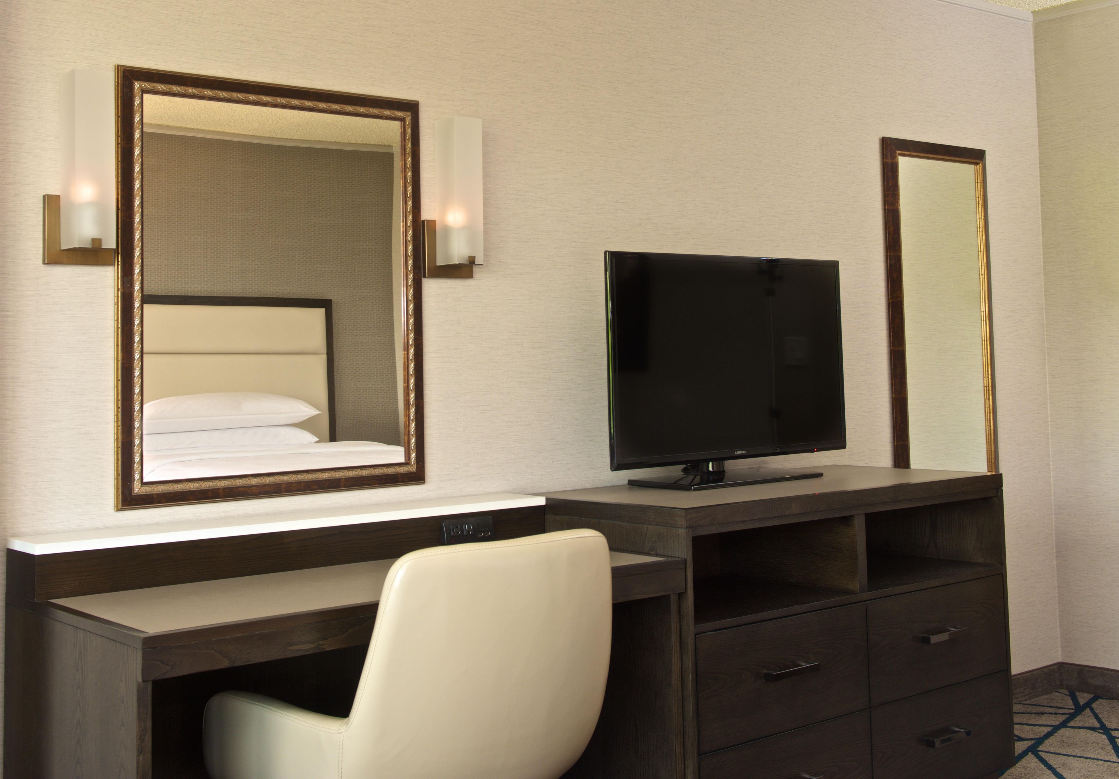 Una habitación de hotel con una cama, televisor, escritorio y silla, y un espejo.