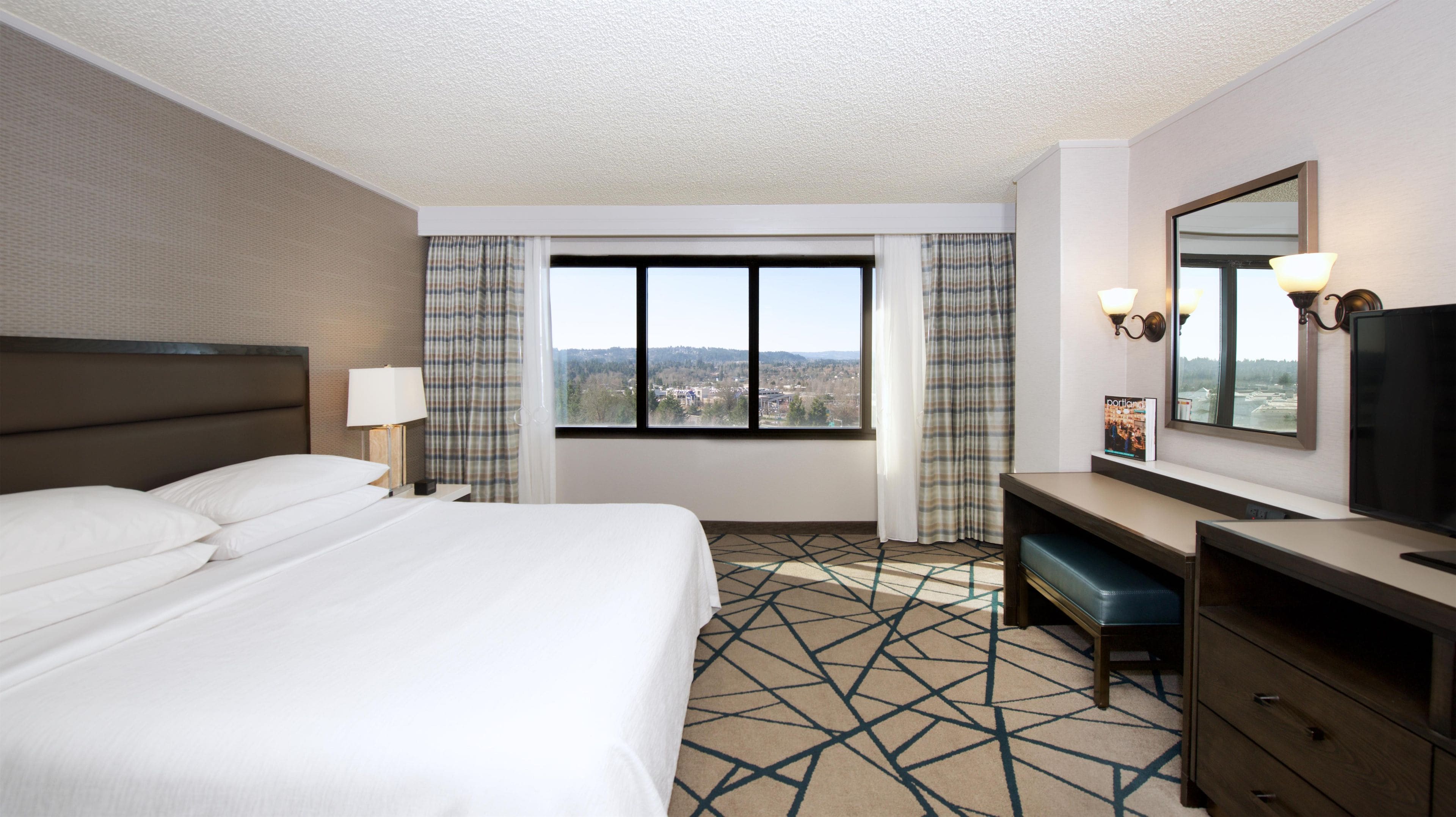 A hotel room with a bed, a desk, a chair, a mirror, and a window with a view of a cityscape.