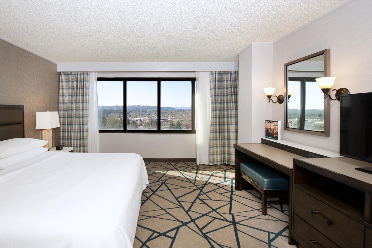 A hotel room with a bed, a desk, a chair, a mirror, and a window with a view of a cityscape.