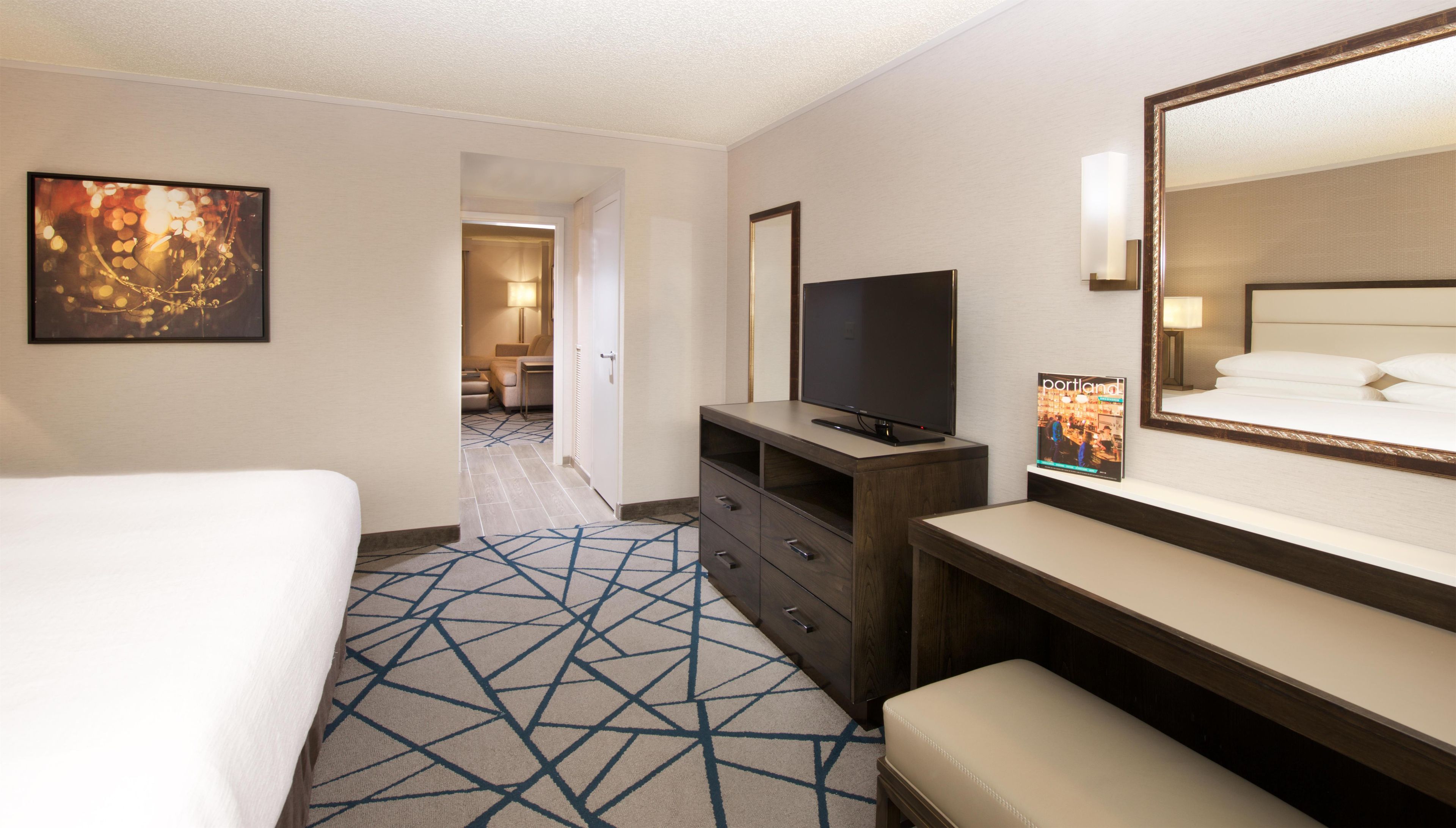 A hotel room with a bed, a television, a dresser, a mirror, and a carpet with a geometric pattern.