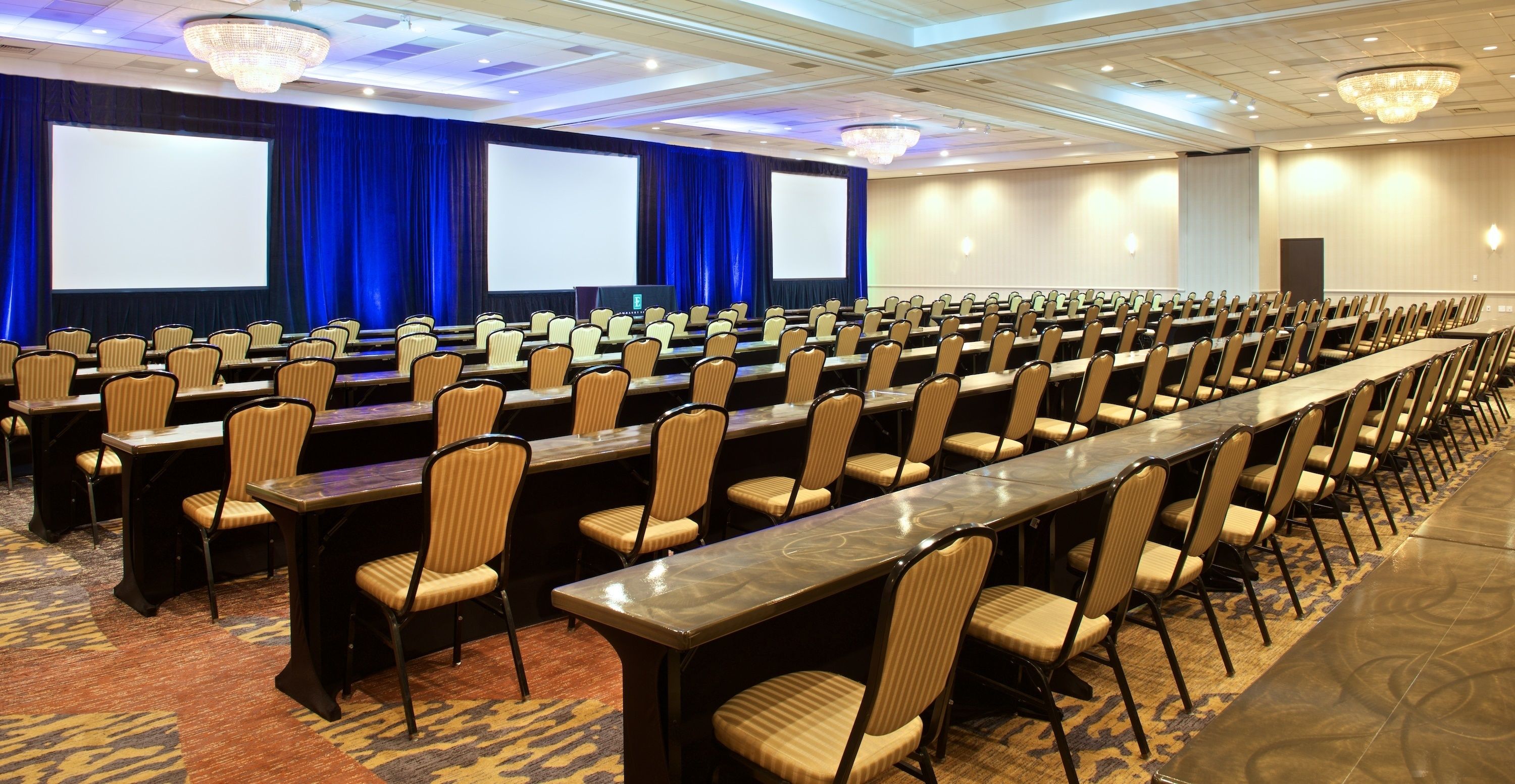 A conference room with rows of chairs and tables, large screens, and chandeliers.