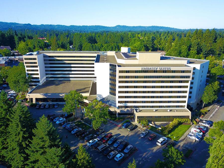 A modern building with a sign reading "Embassy Suites." It features a parking lot with several cars and is surrounded by trees.