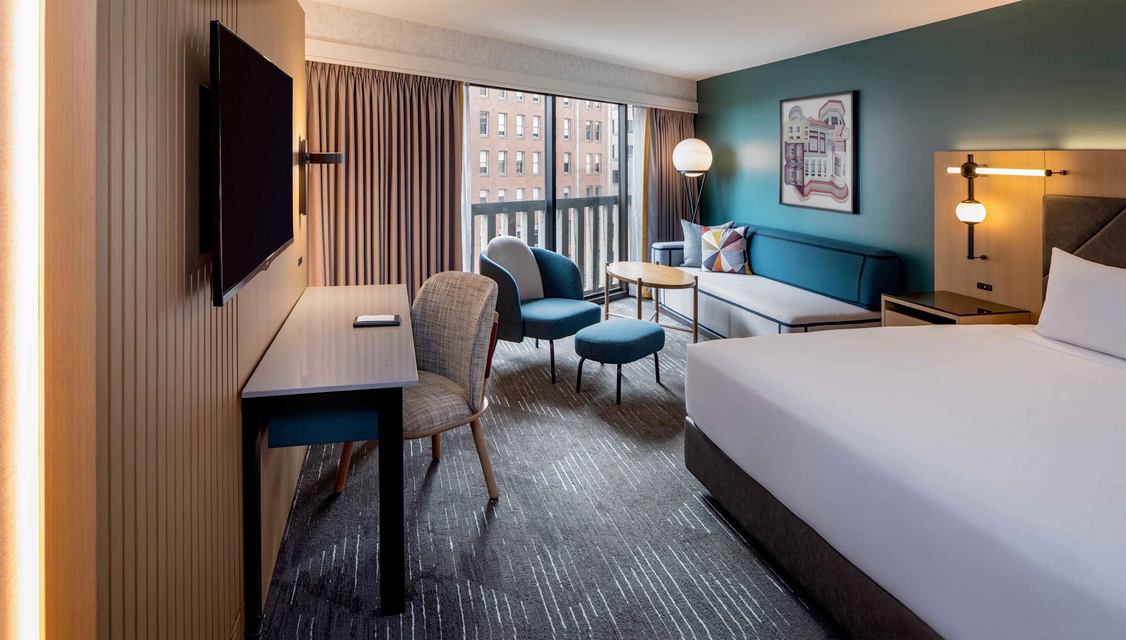 A hotel room with a bed, a desk, a chair, a sofa, and a view of buildings through large windows.