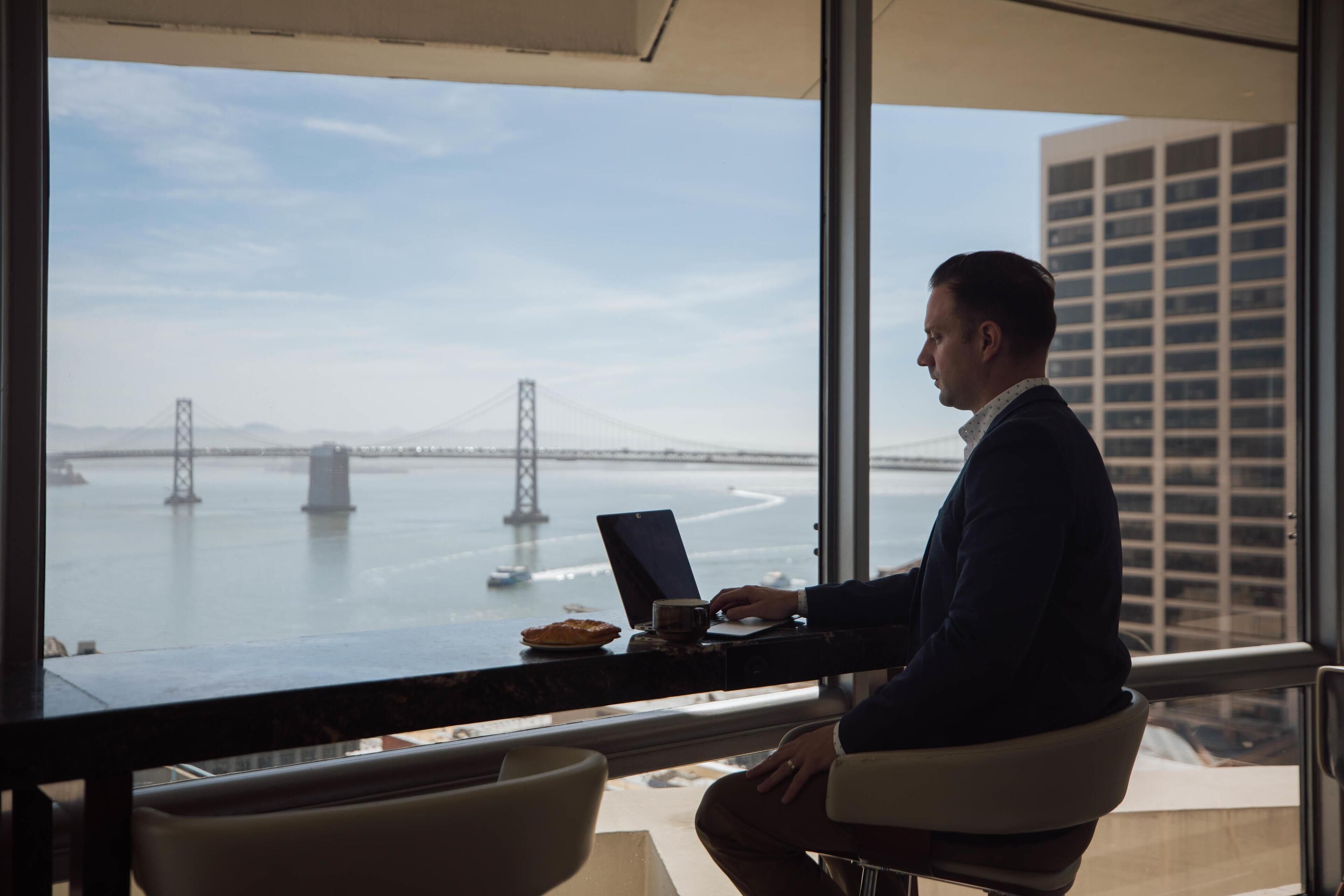 A man working at a desk by a window with a view of a bridge and water.
