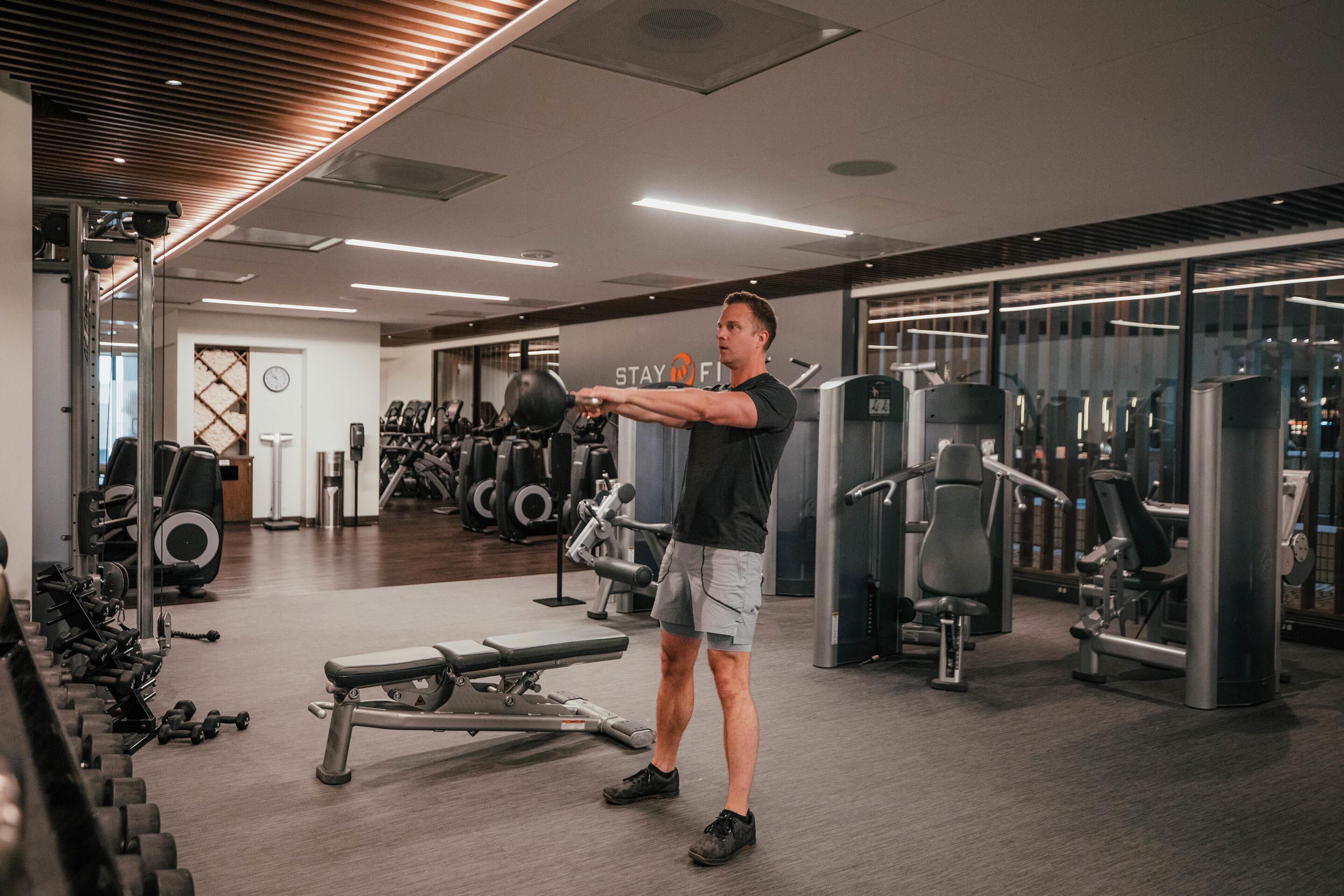 A man exercising with dumbbells in a gym with various fitness equipment.