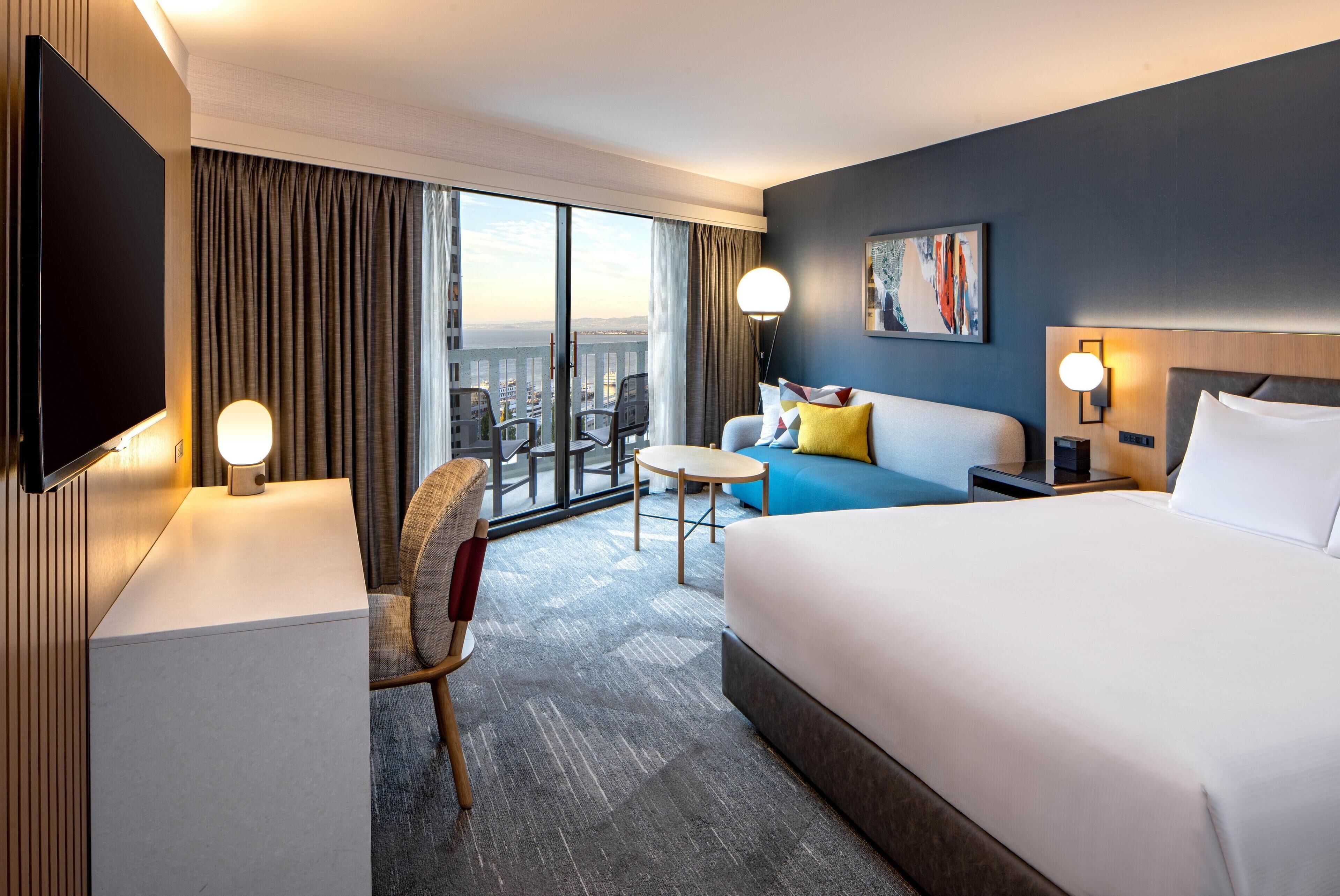 A hotel room with a large bed, a desk, a chair, a small table, a TV, and a balcony with a view.