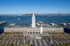 A large building with a prominent clock tower, a spacious plaza with seating, and a view of the water and bridges in the background.