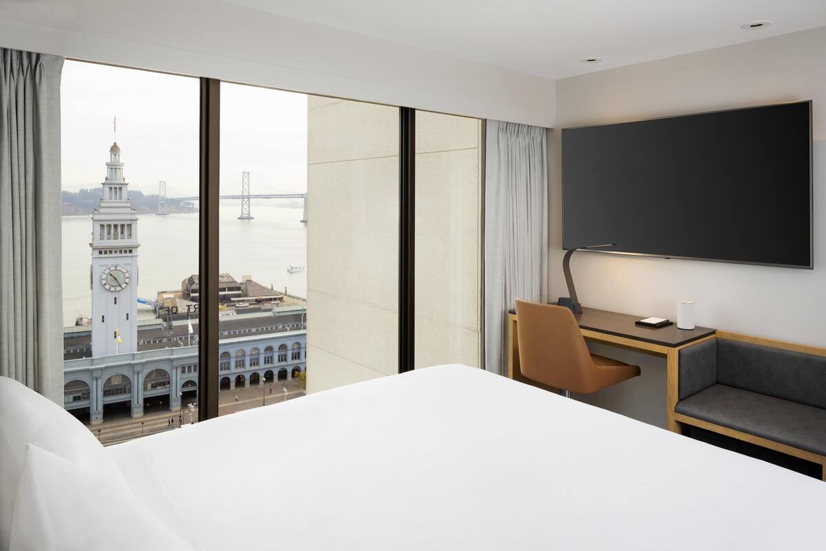 Camera d'albergo con un letto grande, una scrivania, una sedia e vista sulla città, con una torre dell'orologio e un ponte.