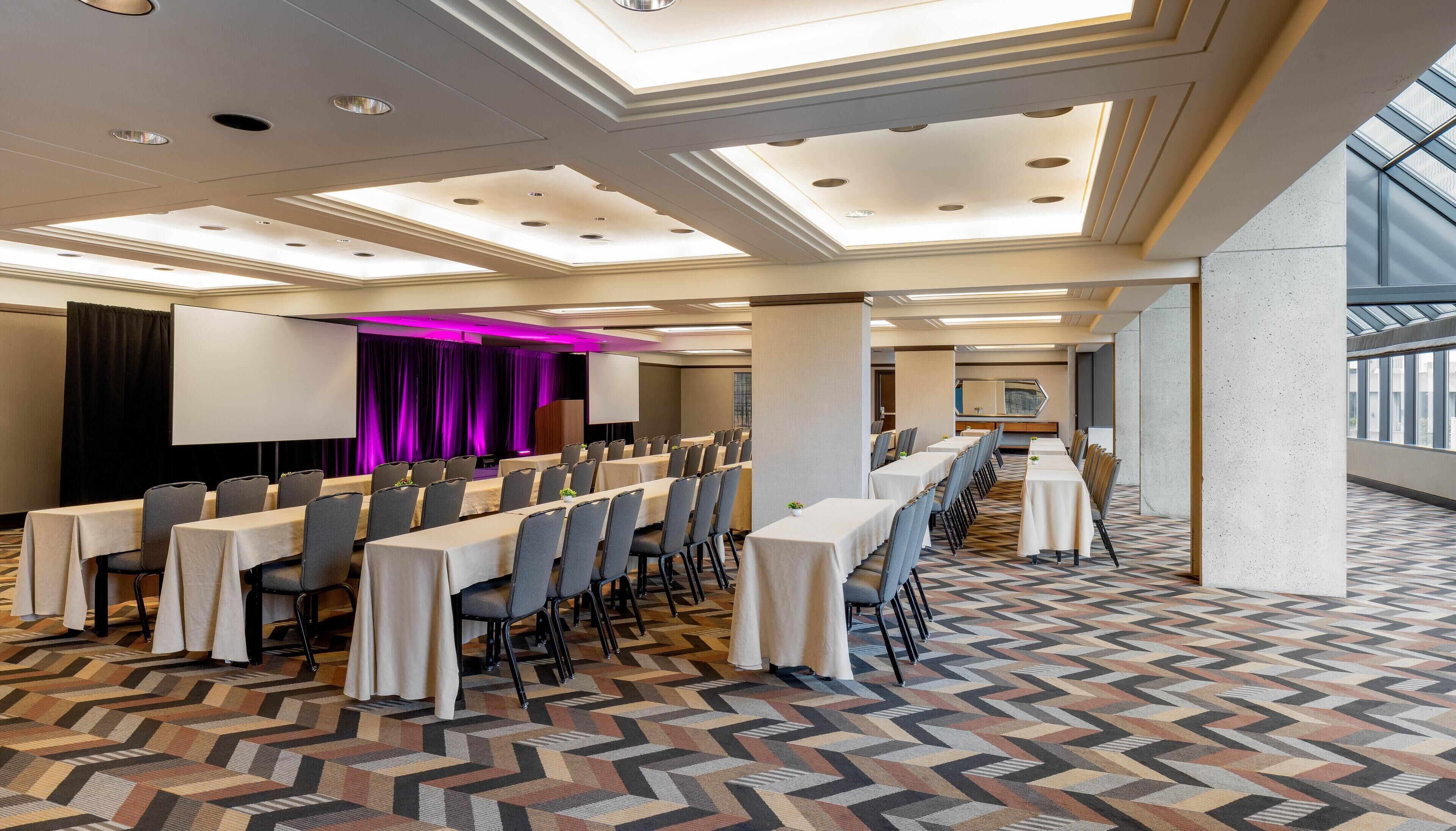 Une salle de conférence équipée de longues tables, de chaises et d’une scène avec un écran.