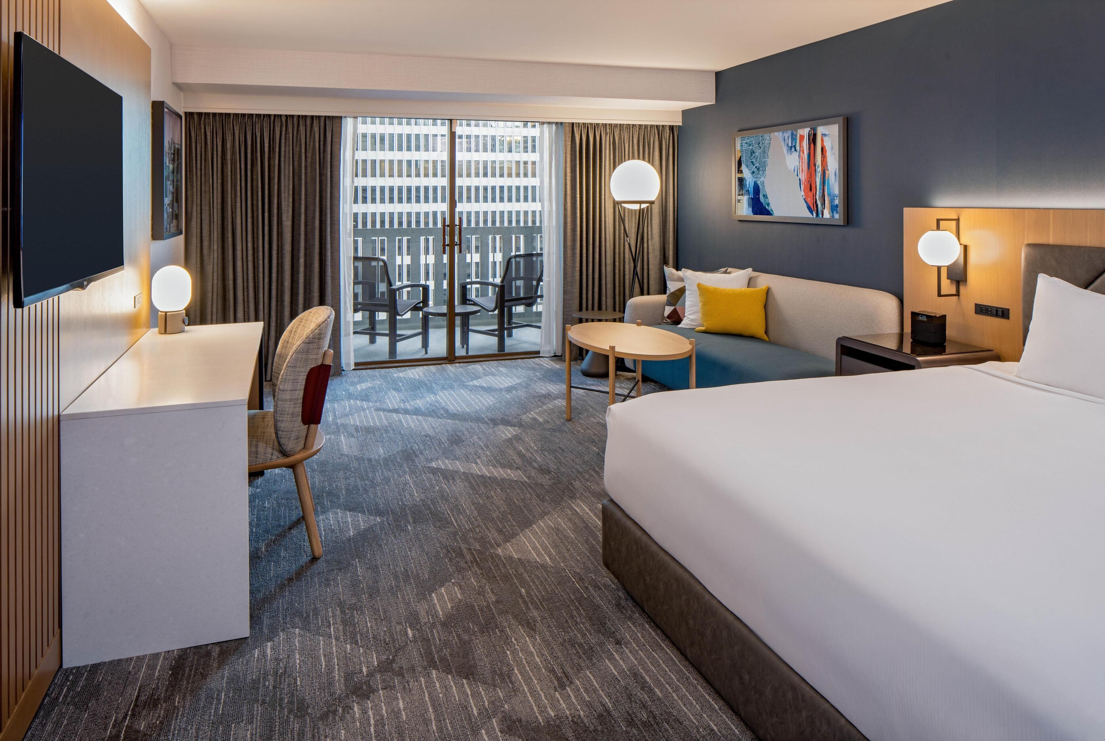 A hotel room with a bed, a desk, a chair, a small table, a sofa, and a TV.