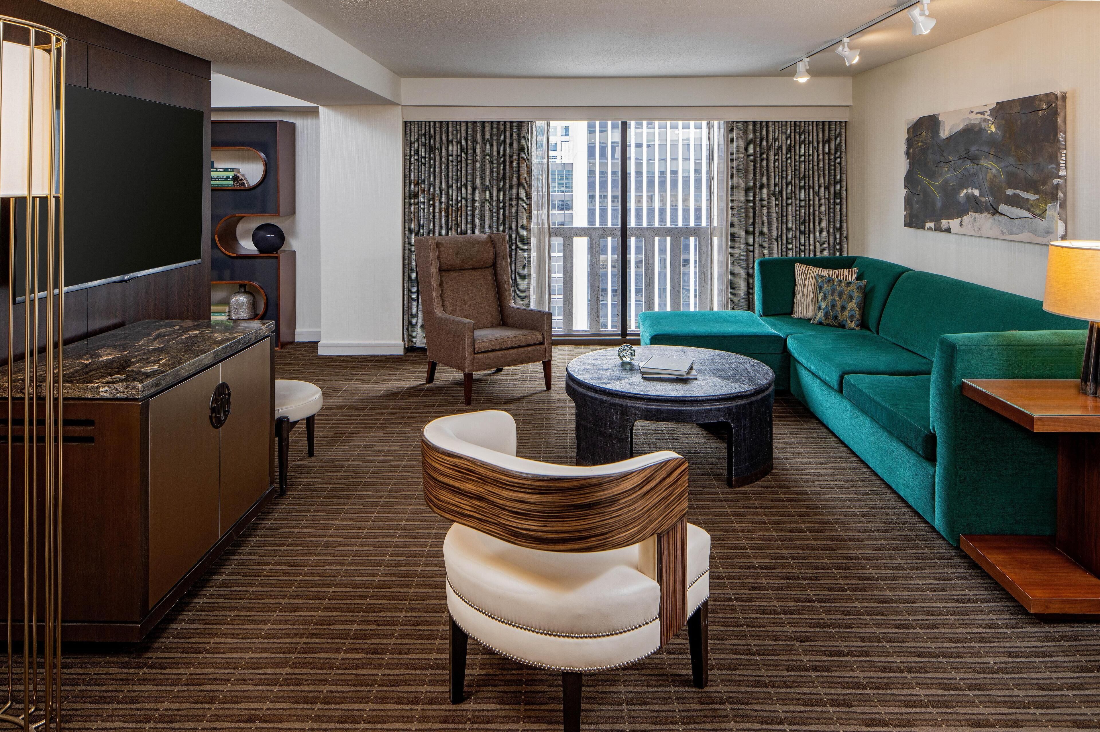 A modern hotel room with a large window, a green sofa, a white armchair, a small round table, and a cabinet with a marble countertop.