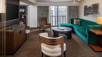 A modern hotel room with a large window, a green sofa, a white armchair, a small round table, and a cabinet with a marble countertop.