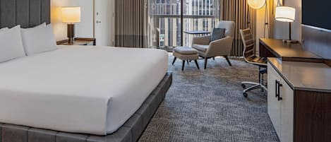 Habitación de hotel con una cama grande, un escritorio, una silla, un televisor y vistas de la ciudad desde la ventana.