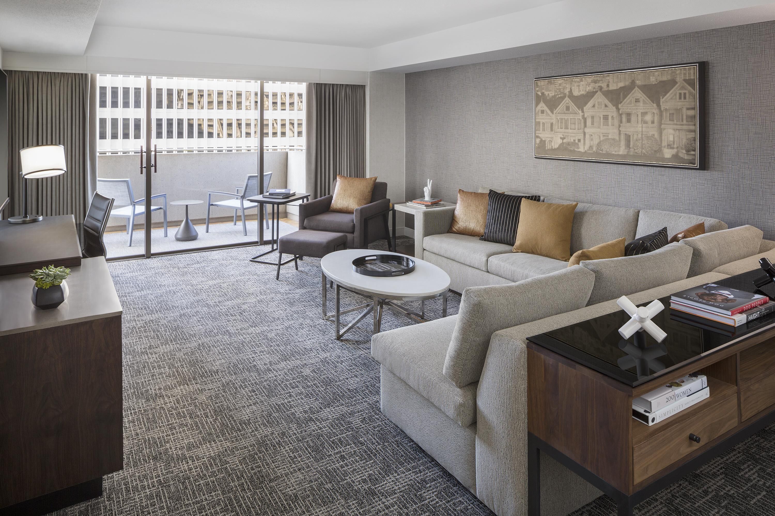 A hotel room with a sofa, armchair, coffee table, and a desk with a lamp.