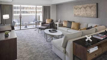 A hotel room with a sofa, armchair, coffee table, and a desk with a lamp.