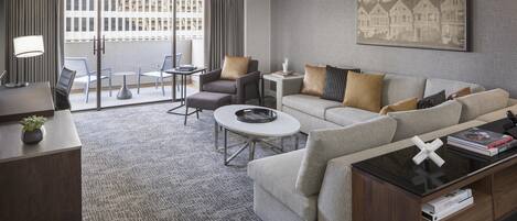 A hotel room with a sofa, armchair, coffee table, and a desk with a lamp.