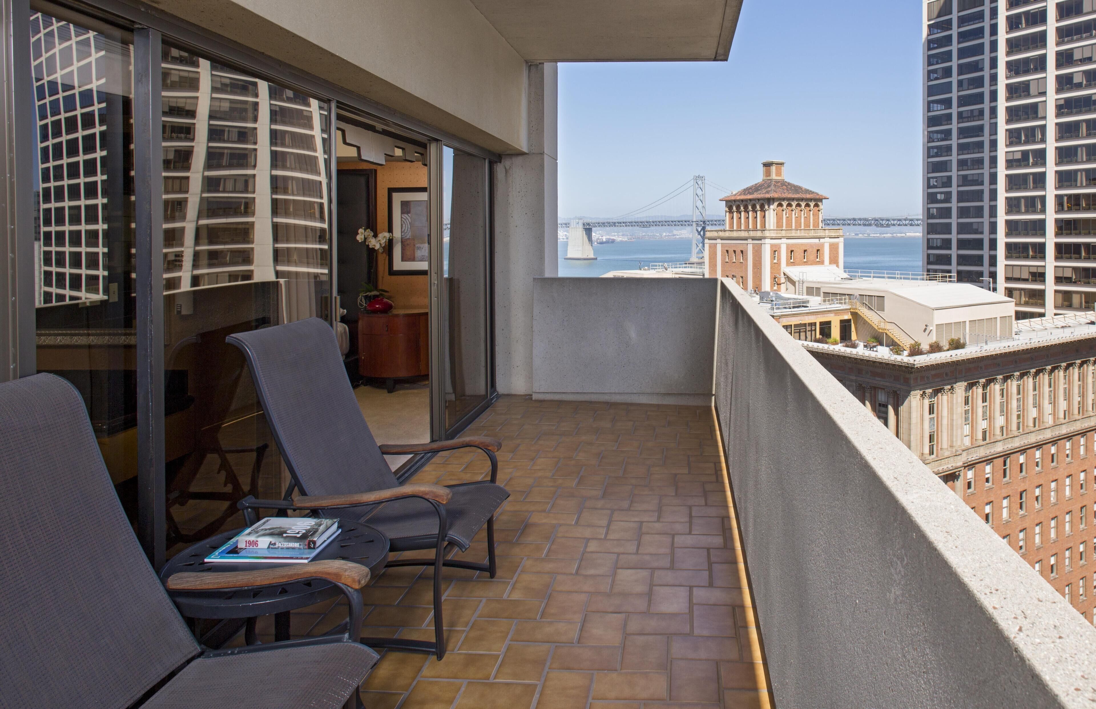 A balcony with two chairs, a small table, and a view of a bridge and buildings.