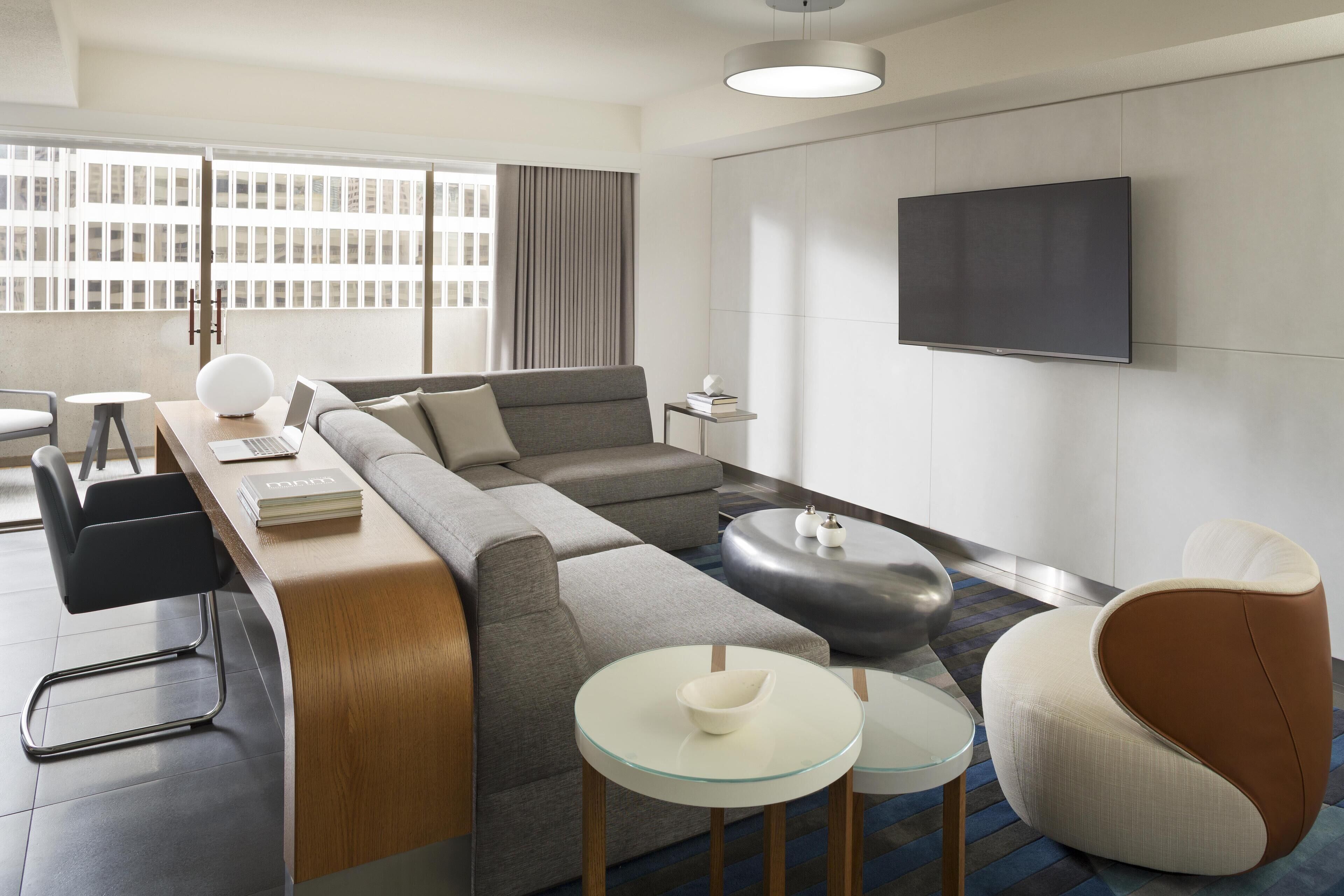 A modern living room with a sectional sofa, a glass coffee table, a round side table, and a flat-screen TV mounted on the wall.