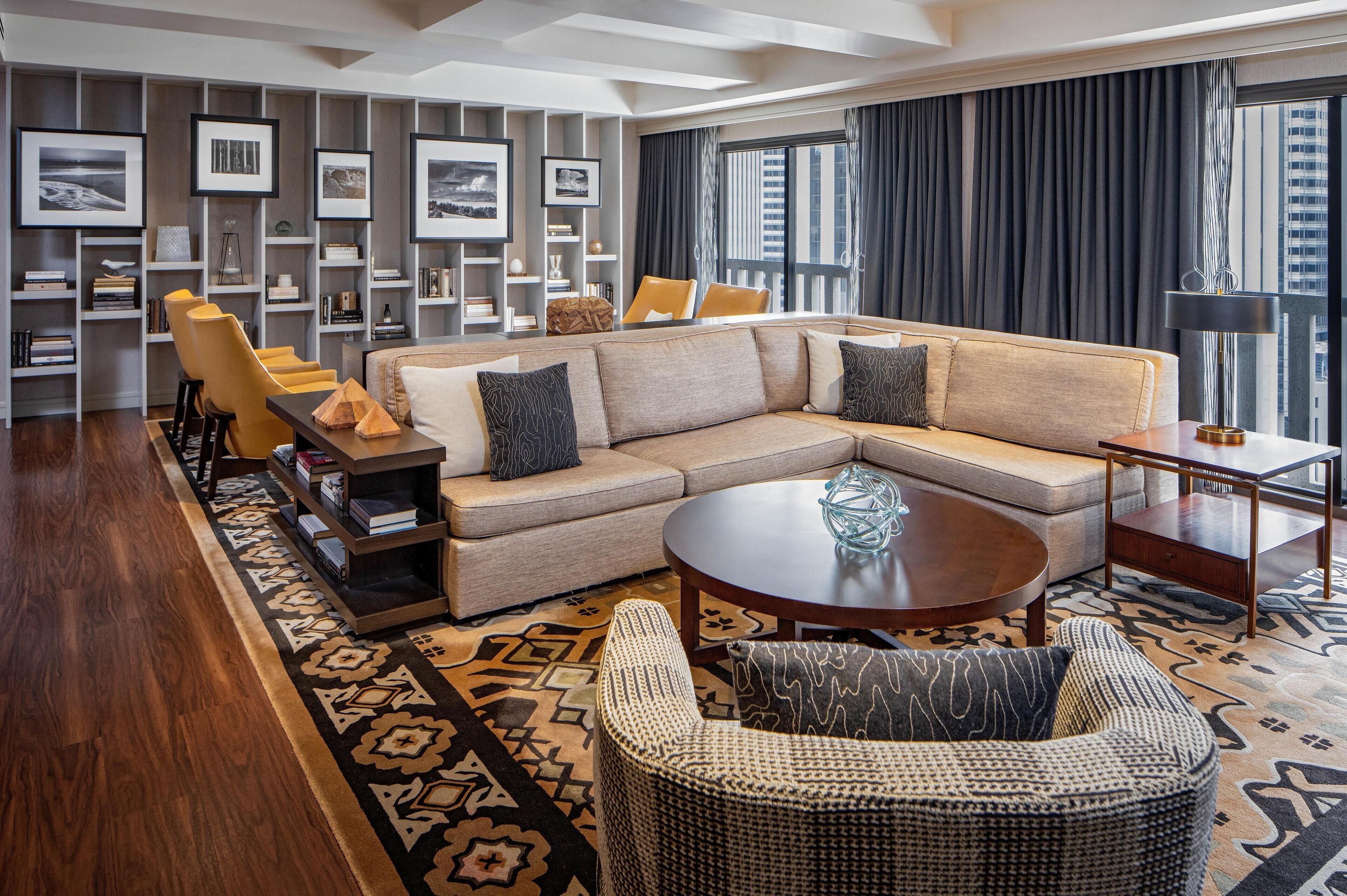 A modern living room with a beige sectional sofa, a round coffee table, and a wicker chair. There are bookshelves with framed pictures and a large window with curtains.
