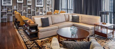 A modern living room with a beige sectional sofa, a round coffee table, and a wicker chair. There are bookshelves with framed pictures and a large window with curtains.