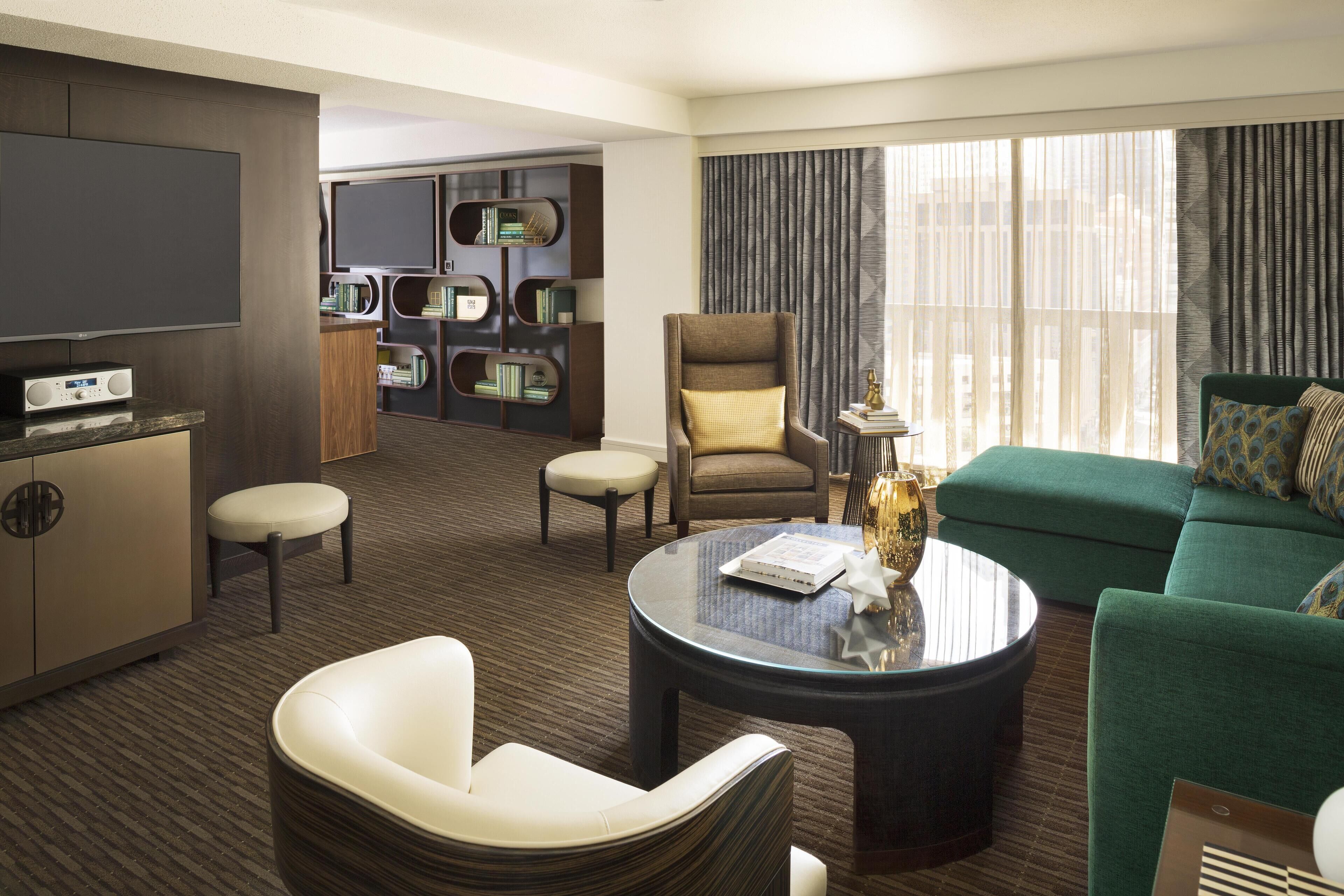A modern hotel room with a green sofa, a round coffee table, a TV, a bookshelf, and a seating area with a chair and ottoman.