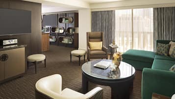 Una habitación de hotel moderna con un sofá verde, una mesa de centro redonda, un televisor, una estantería y una zona de estar con una silla y un reposapiés.