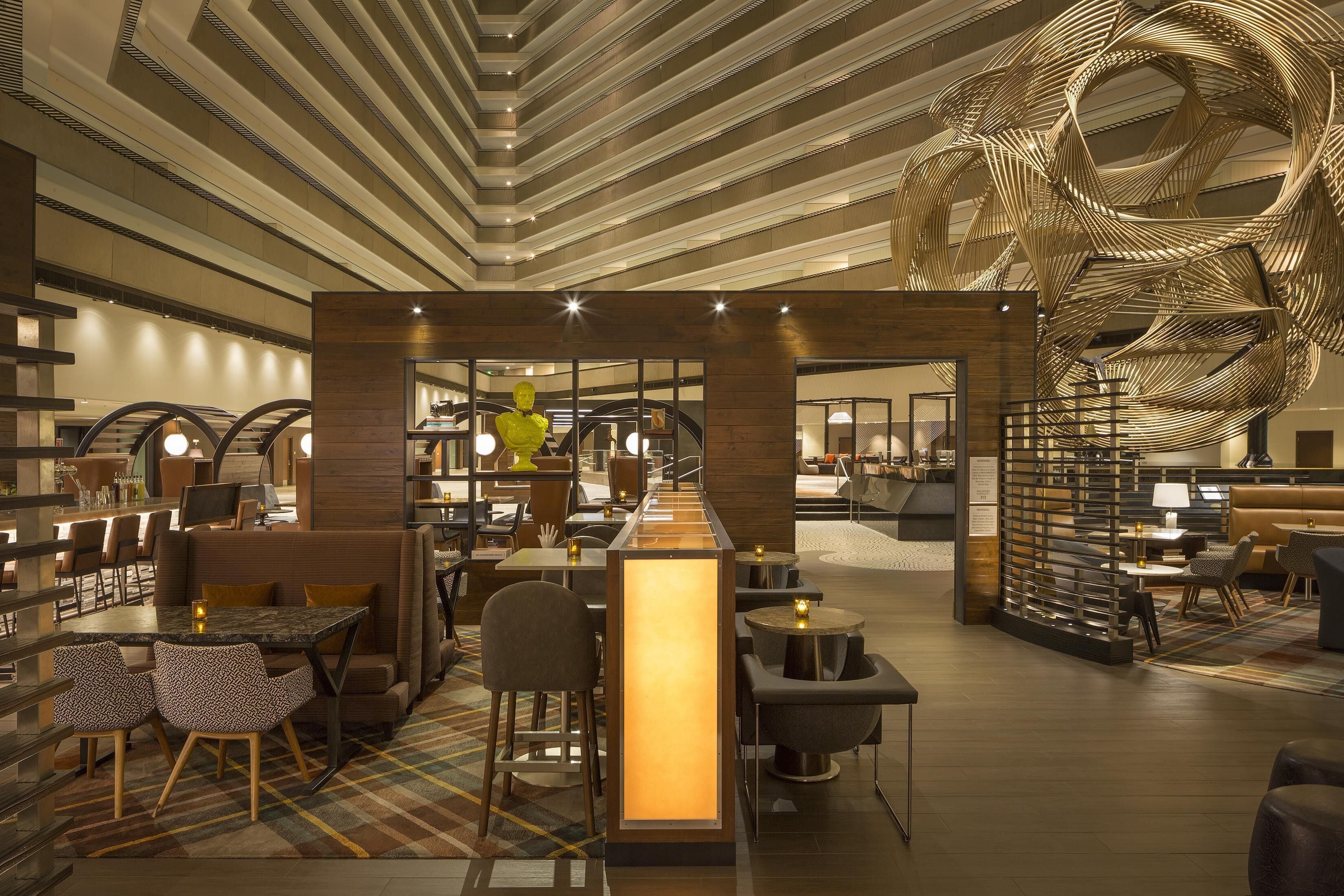 A modern hotel lobby with a central bar, seating areas, and a distinctive ceiling sculpture.