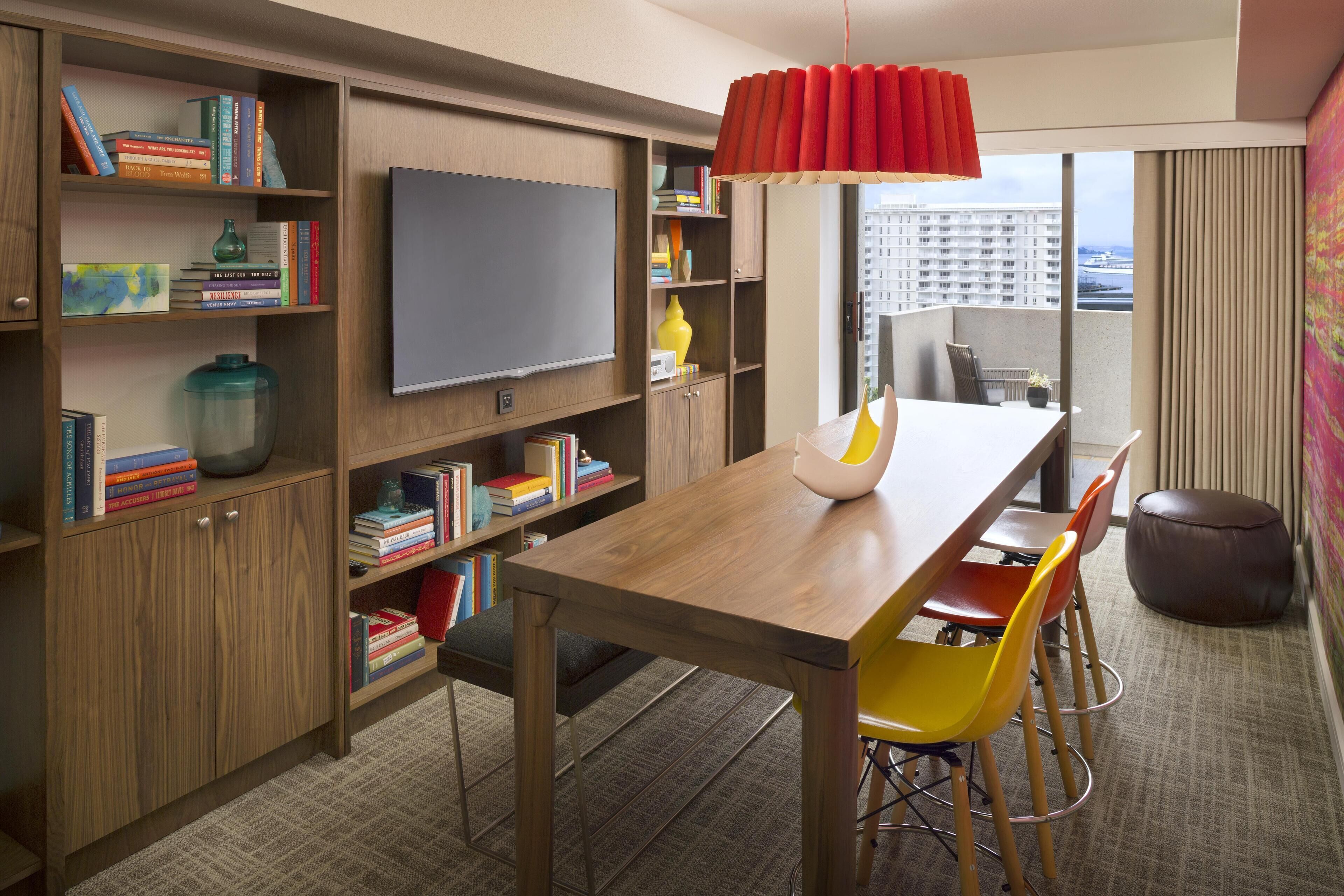 Een moderne hotelkamer met een houten boekenkast, een flatscreen televisie, een eettafel met kleurrijke stoelen en uitzicht op de stad door grote ramen.
