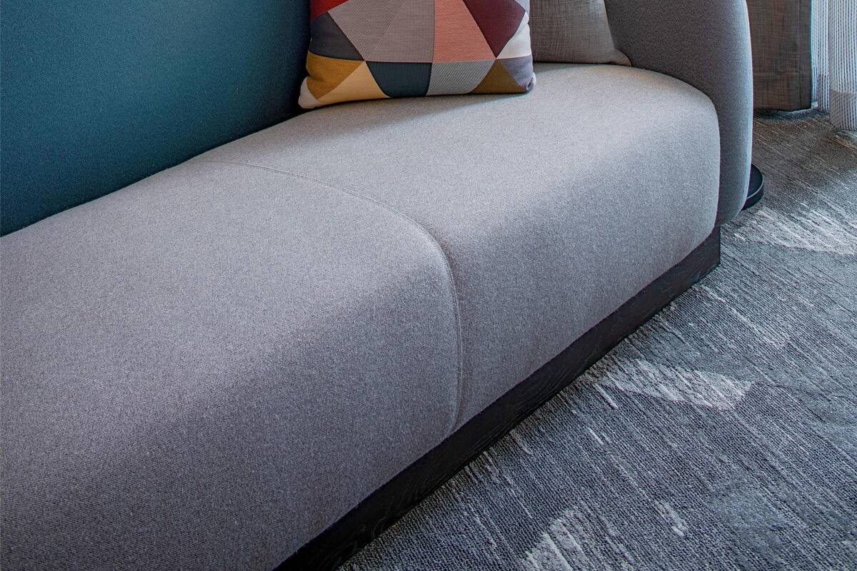 Una camera d'albergo moderna con un divano grigio, un cuscino geometrico colorato, una lampada da terra e un quadro astratto incorniciato appeso alla parete.