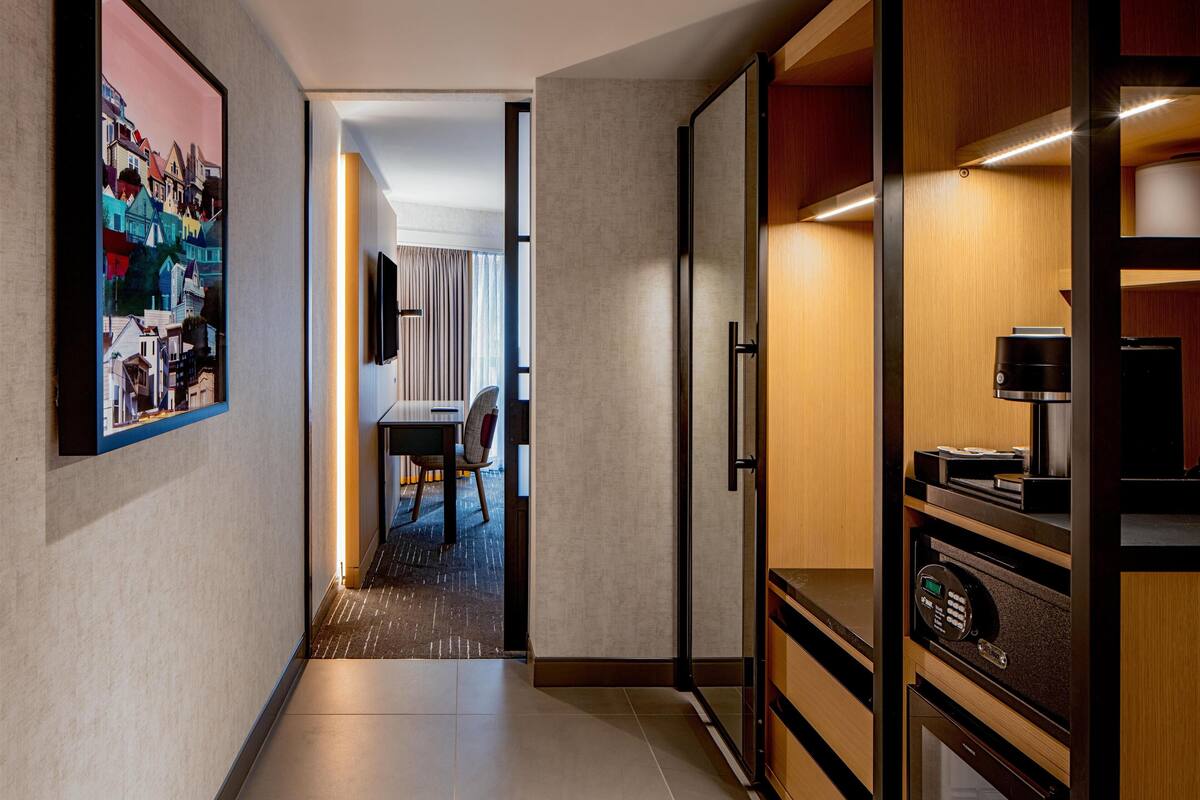 Un corridoio d'hotel con un quadro incorniciato a sinistra, una zona pranzo con tavolo e sedie al centro e un angolo bar con una macchina da caffè a destra.