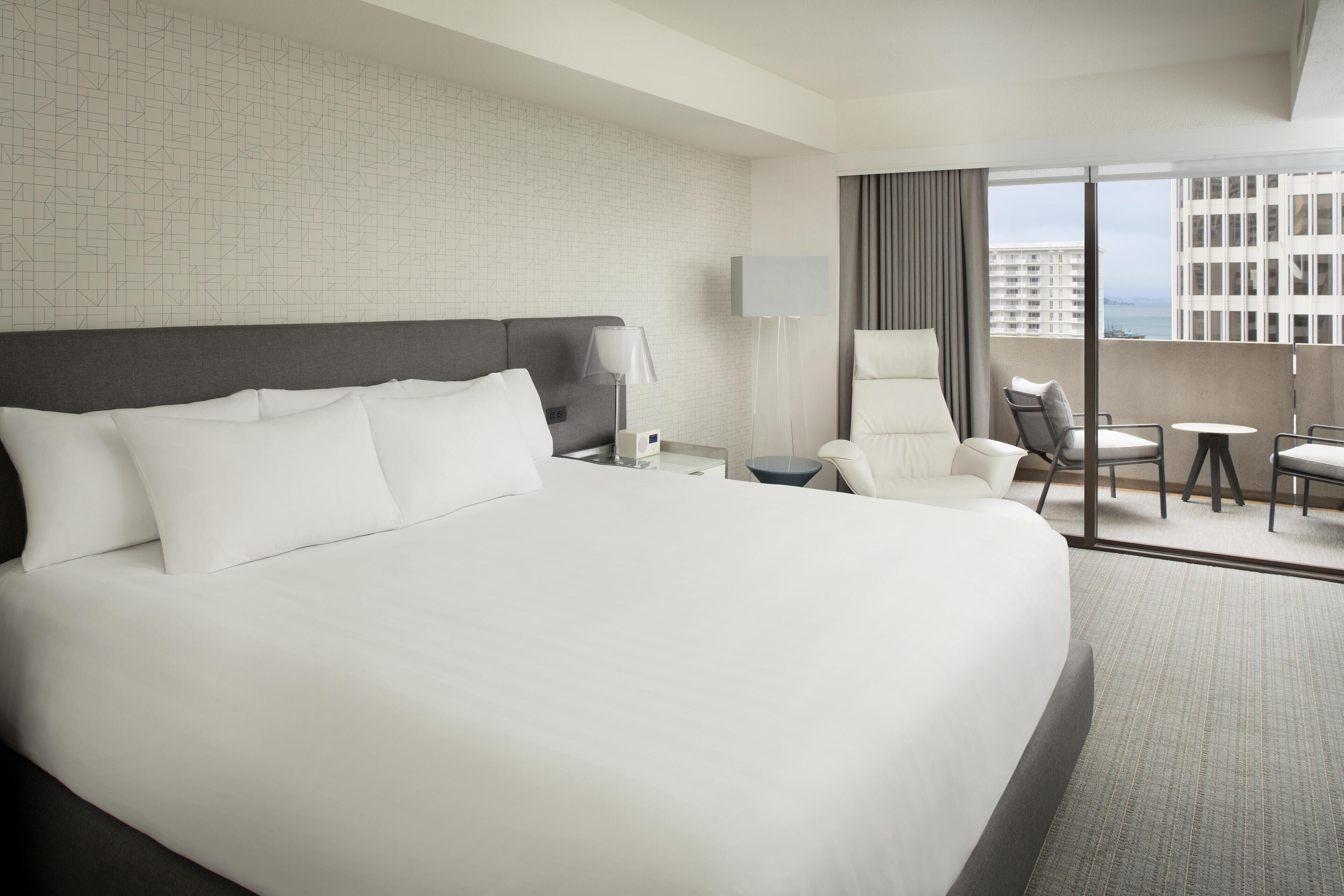A modern hotel room with a large bed, a desk, a chair, and a view of the city.
