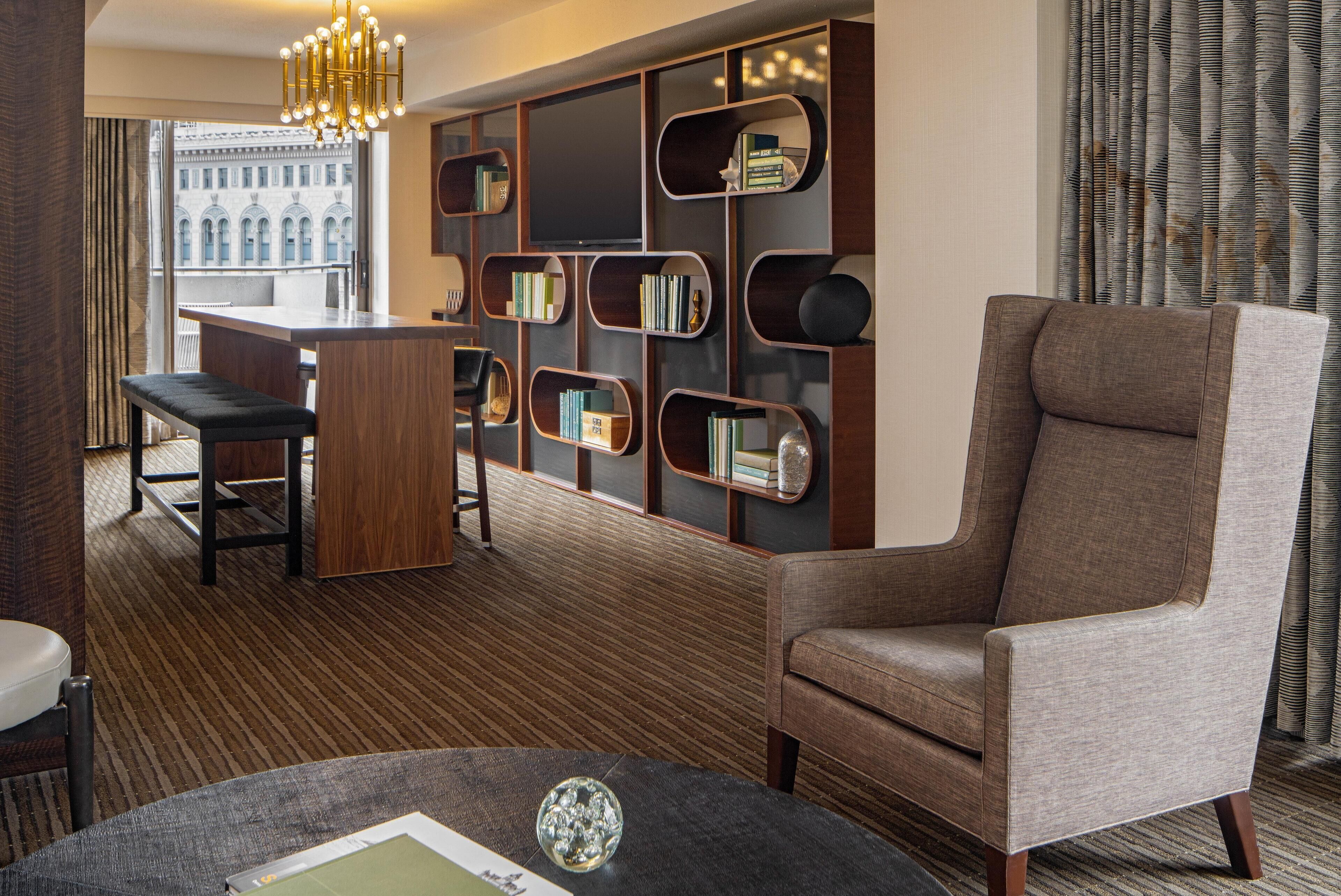 Una habitación de hotel moderna con un ventanal grande, una zona de bar, un espacio de descanso con sofá y sillas, y una estantería con libros.