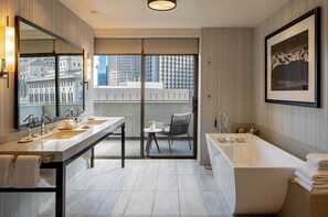 Een moderne badkamer met een groot bad, twee wasbakken en een grote spiegel. Er staat een stoel en een klein tafeltje in de hoek, en een balkon met uitzicht op gebouwen.