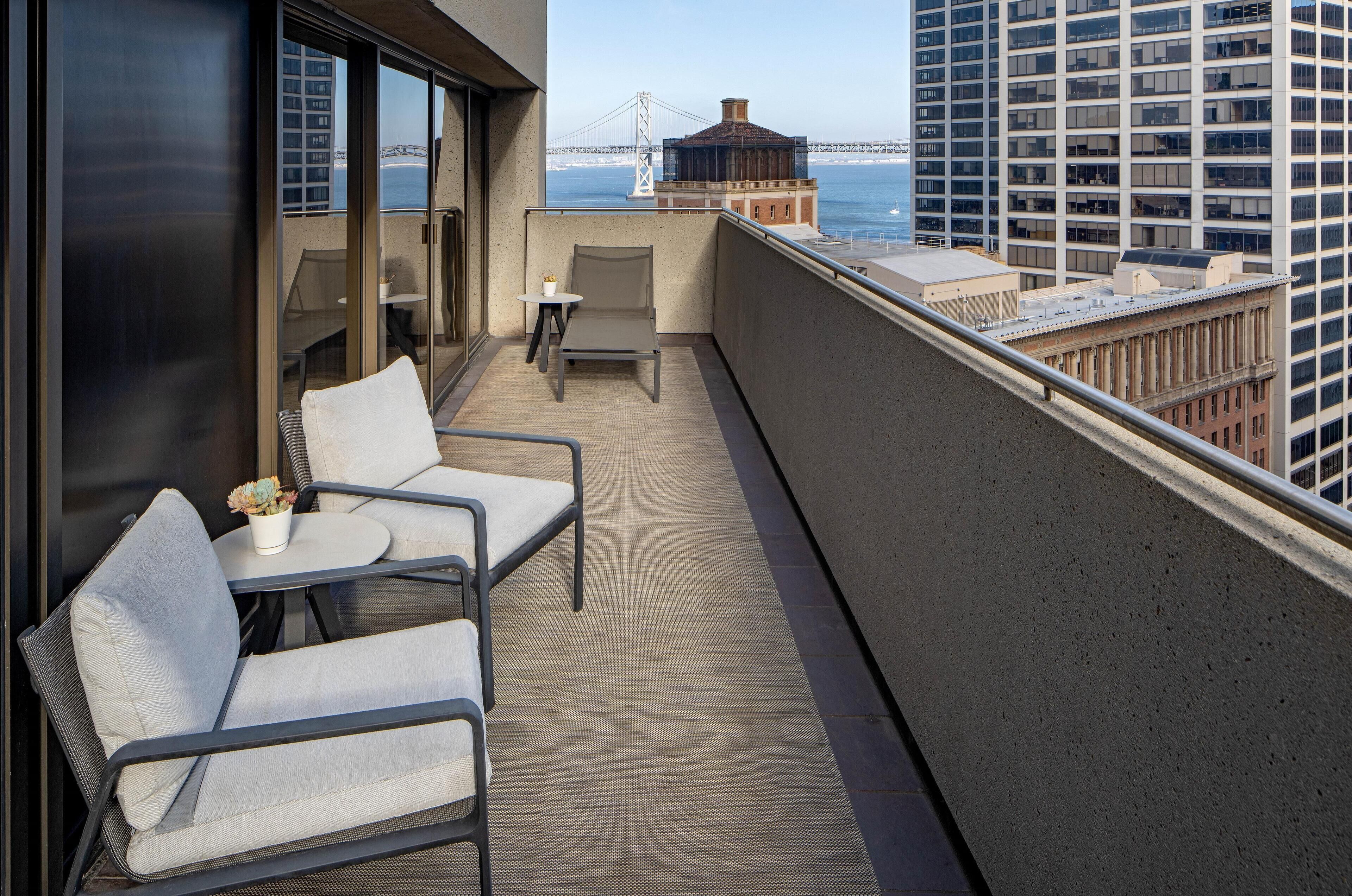A balcony with two chairs and a small table, overlooking a cityscape and a bridge.