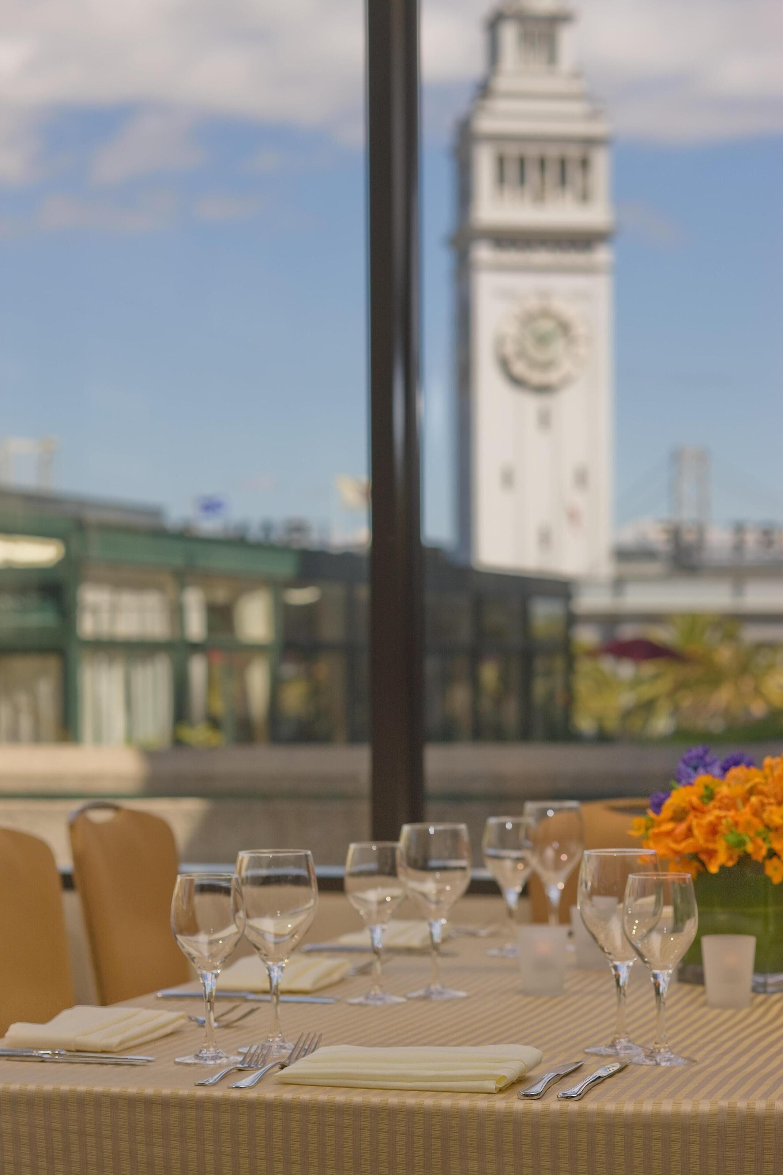 A dining table set with wine glasses, cutlery, and a vase of flowers, with a view of a clock tower in the background.