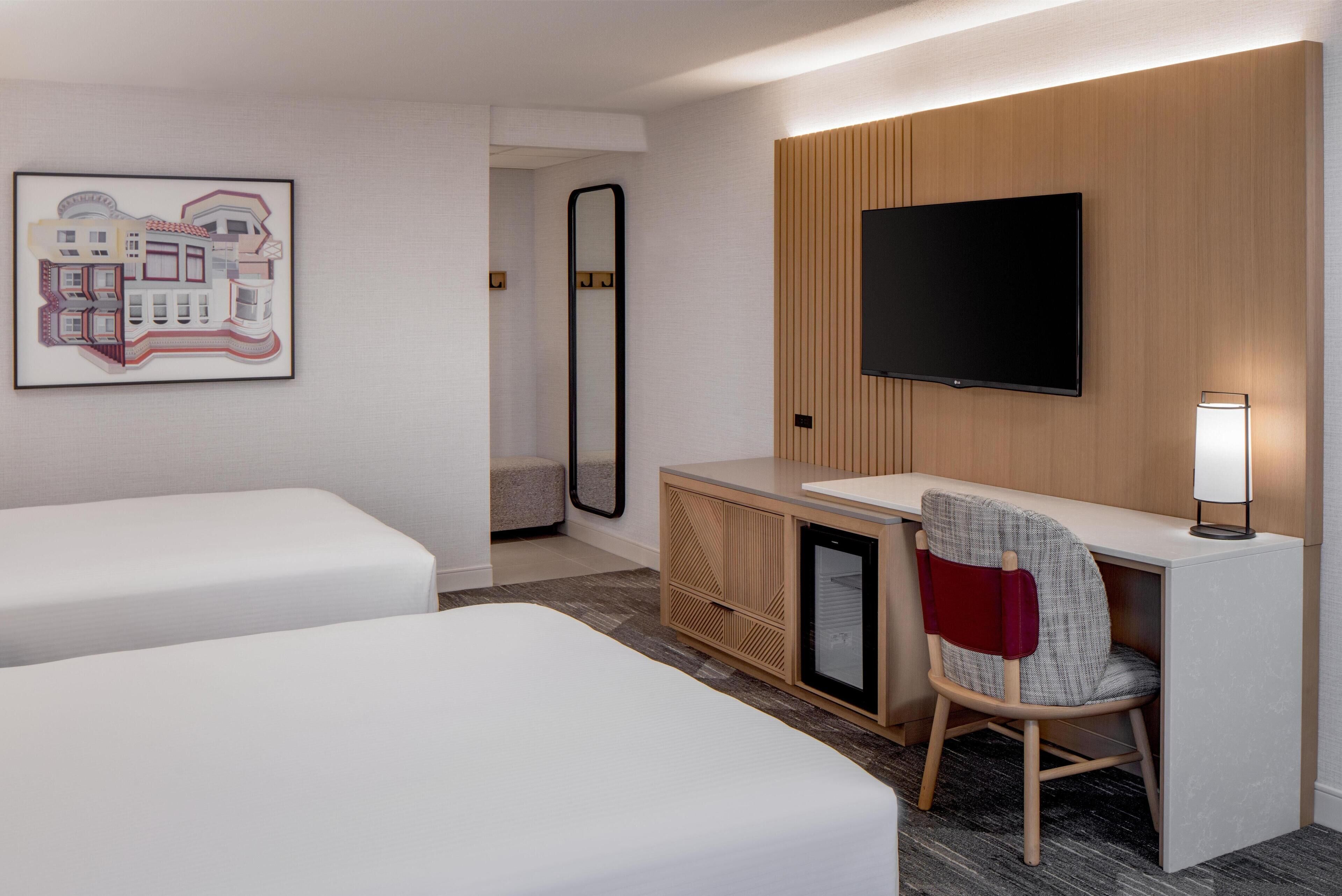 A hotel room with two beds, a desk with a chair, a flat-screen TV, a mirror, and a framed artwork on the wall.