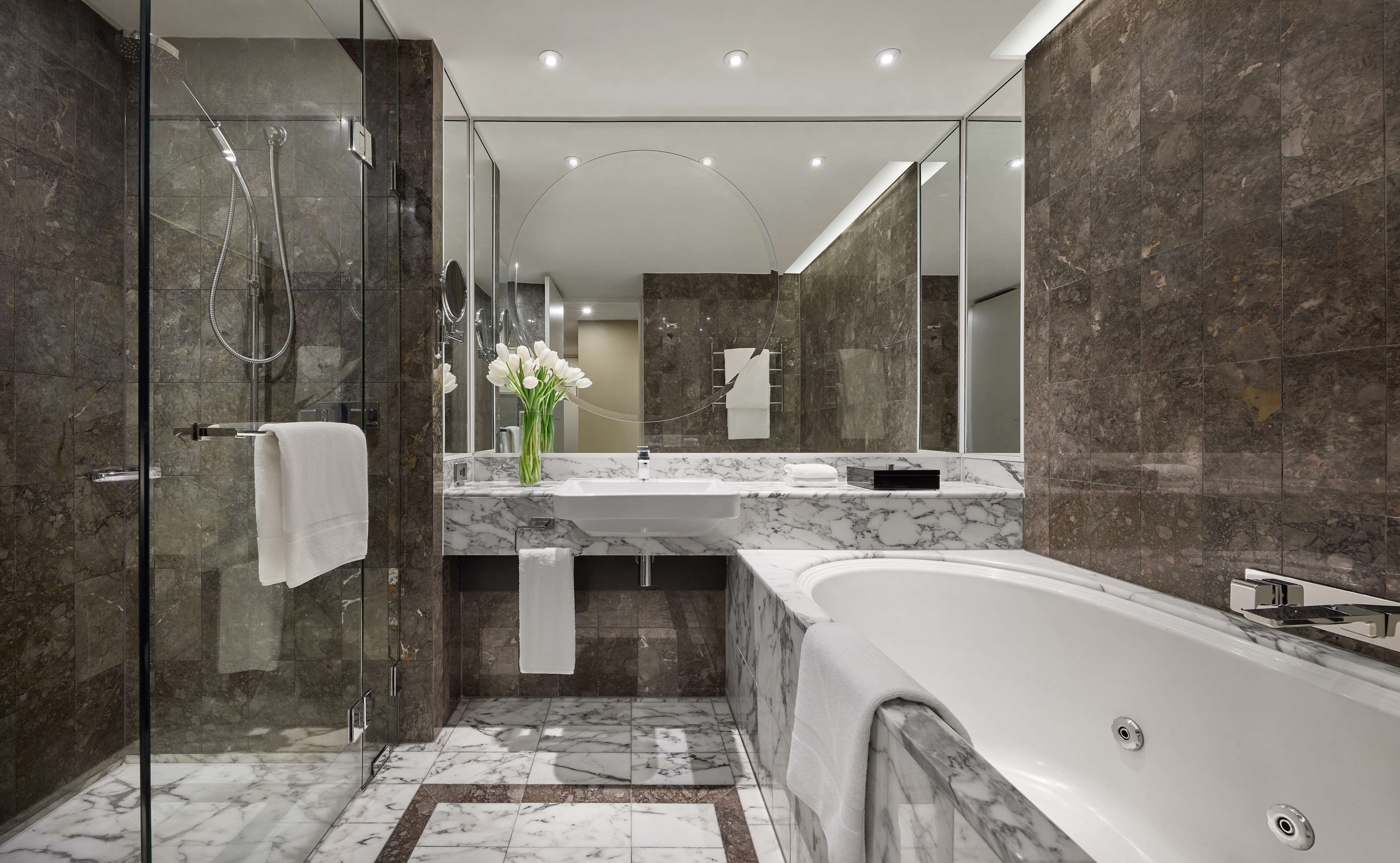 A modern bathroom with a glass-enclosed shower, a marble-tiled floor, a large bathtub, and a vanity with a mirror.