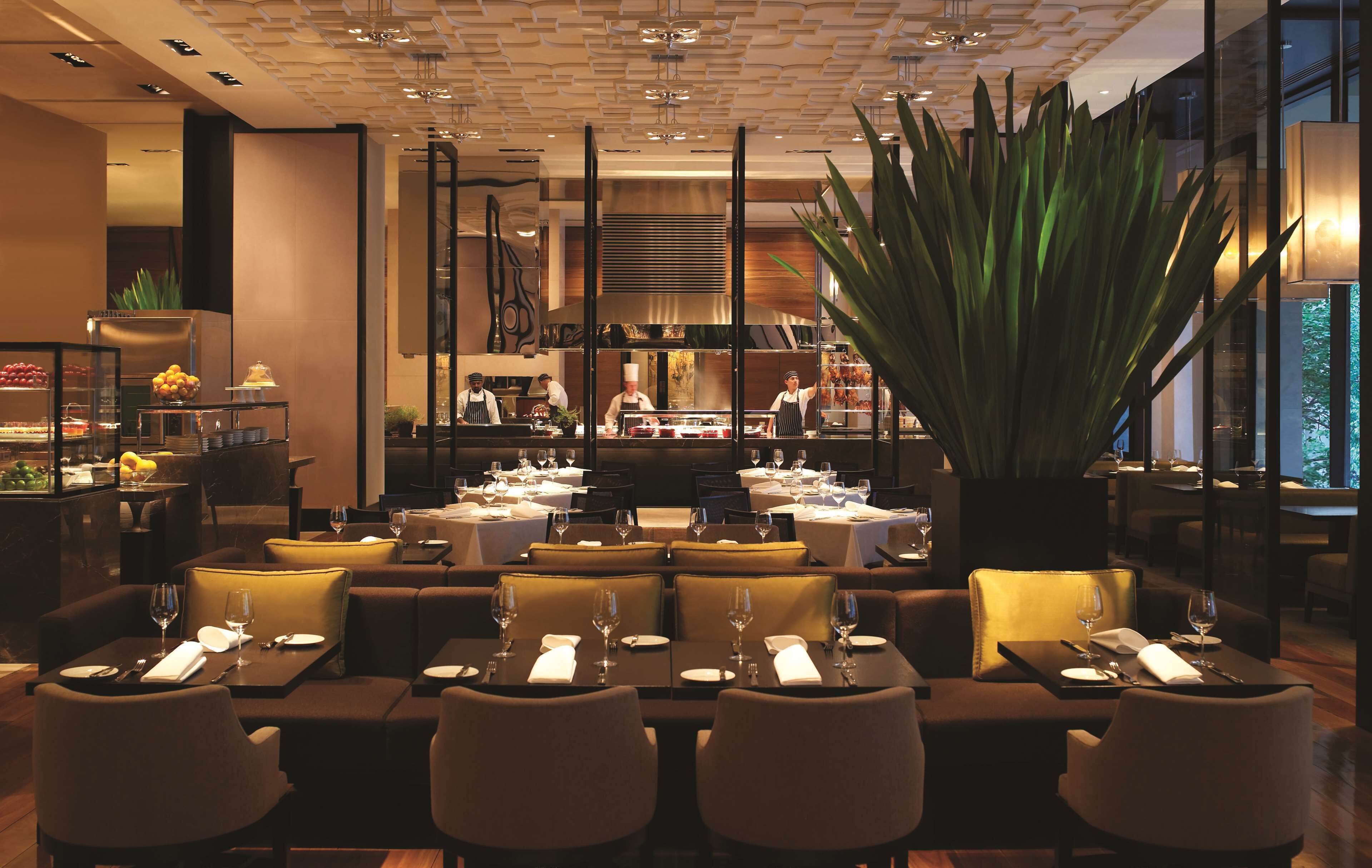A restaurant with tables set for dining, a large plant in the center, and a kitchen area in the background.