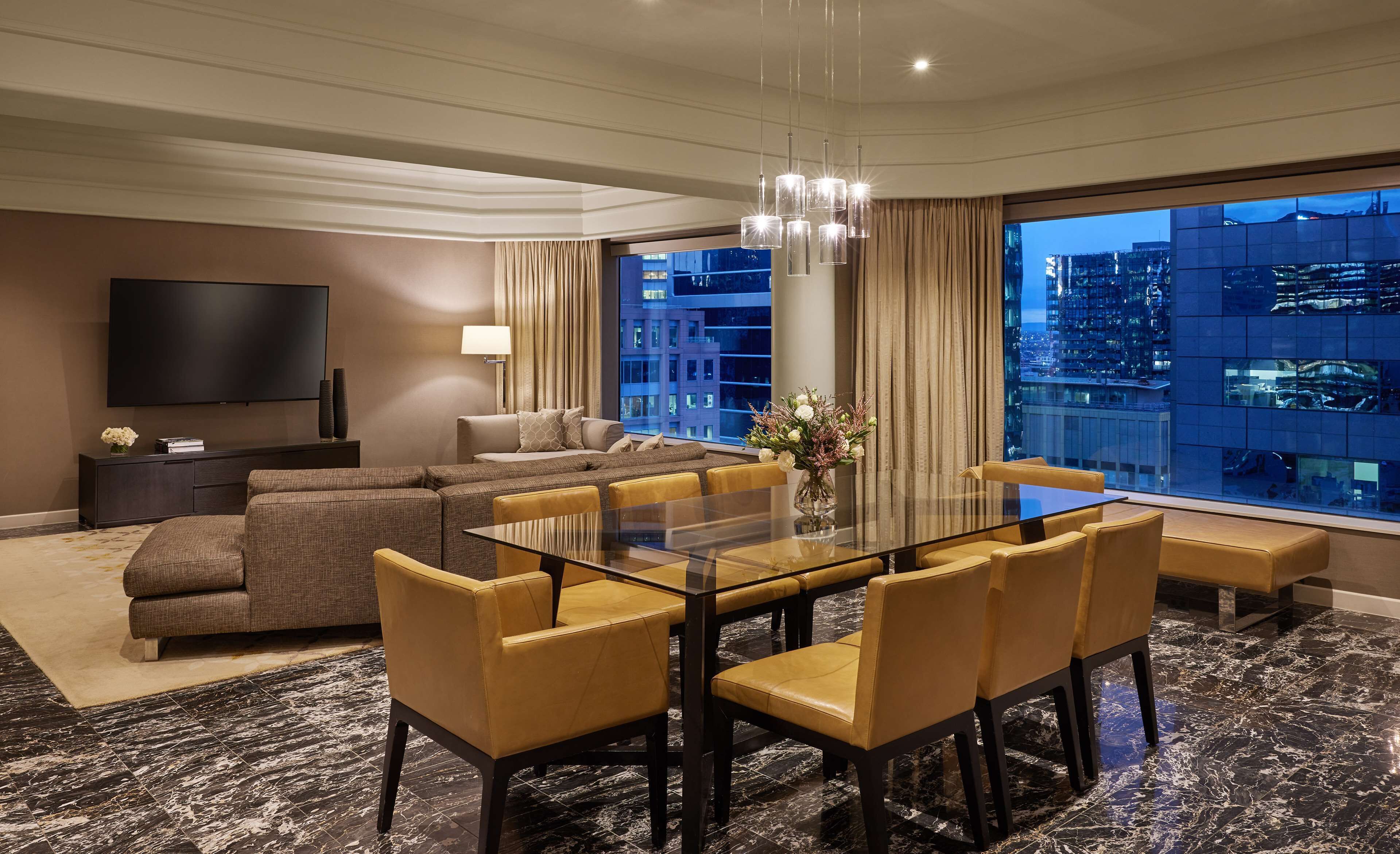 A modern hotel room with a large dining table, comfortable seating, a flat-screen TV, and a city view.