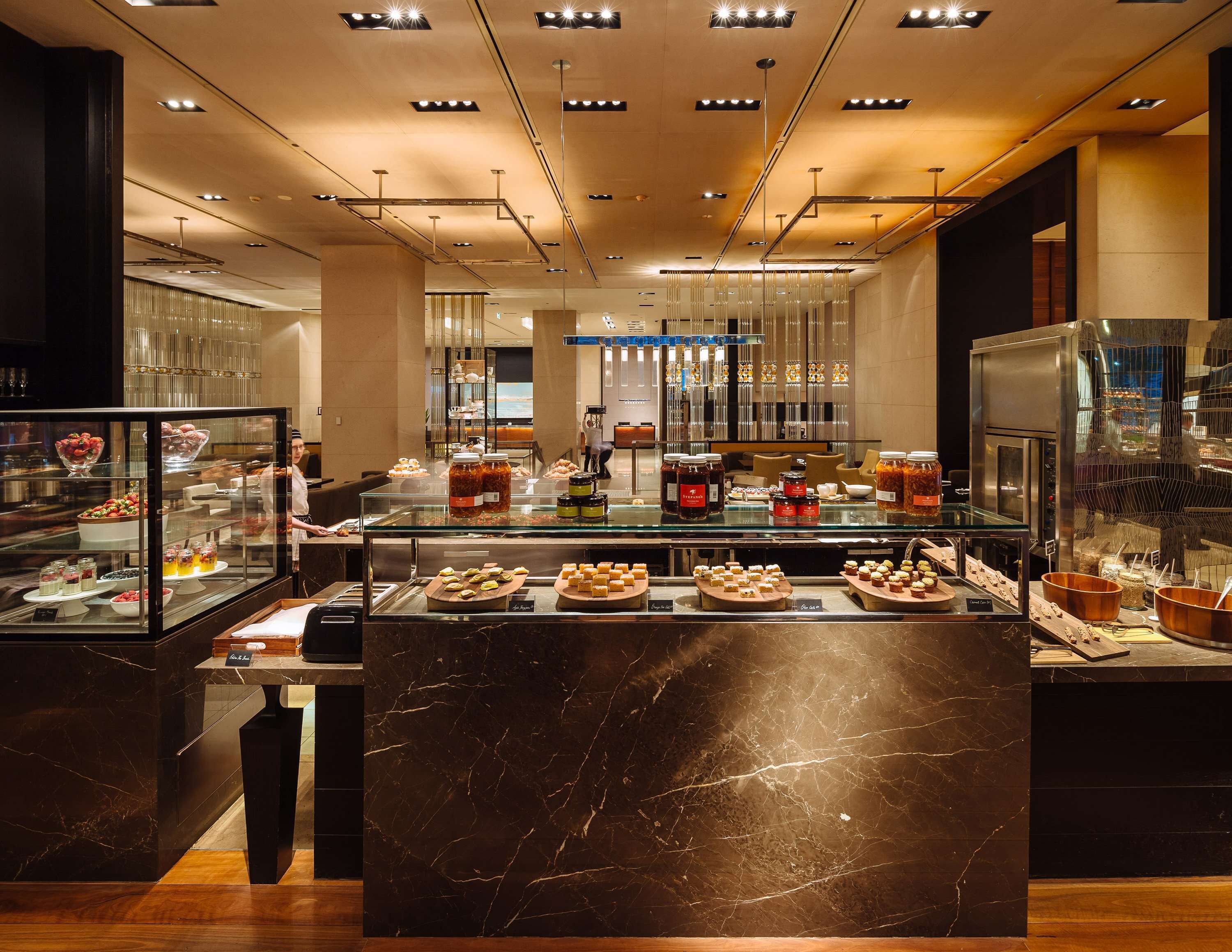 A modern hotel lobby with a marble reception desk, a glass display case with pastries, and a seating area with a view of the cityscape.
