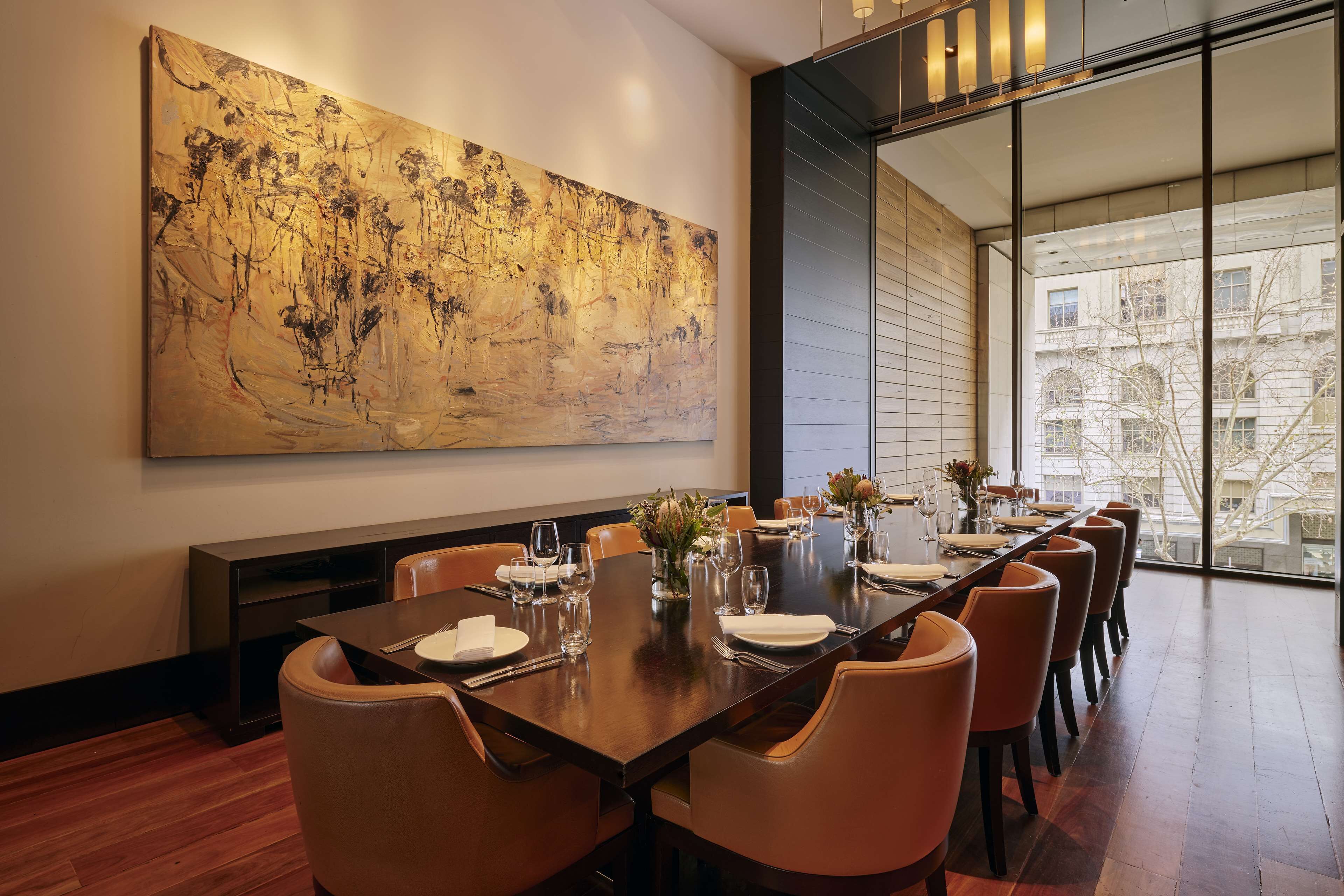 Ein Esszimmer mit einem langen Tisch, gedeckt für eine Mahlzeit, einem großen abstrakten Gemälde an der Wand und Blick auf Gebäude durch große Fenster.